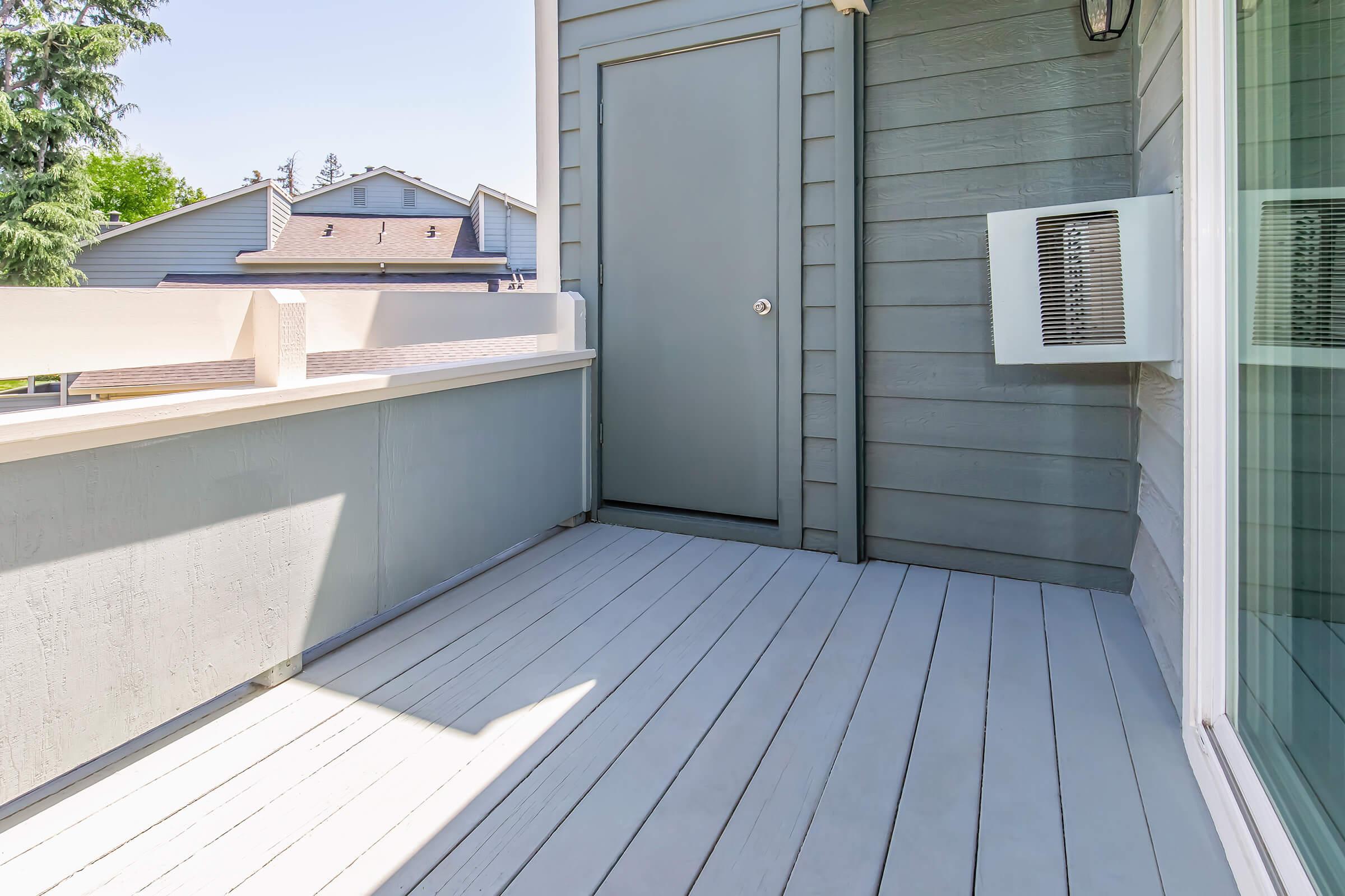 A clean, modern balcony space with light gray flooring and a wall-mounted air conditioning unit. There is a closed door leading inside, and a view of rooftops and trees in the background under a clear blue sky. The space is well-lit by natural sunlight.
