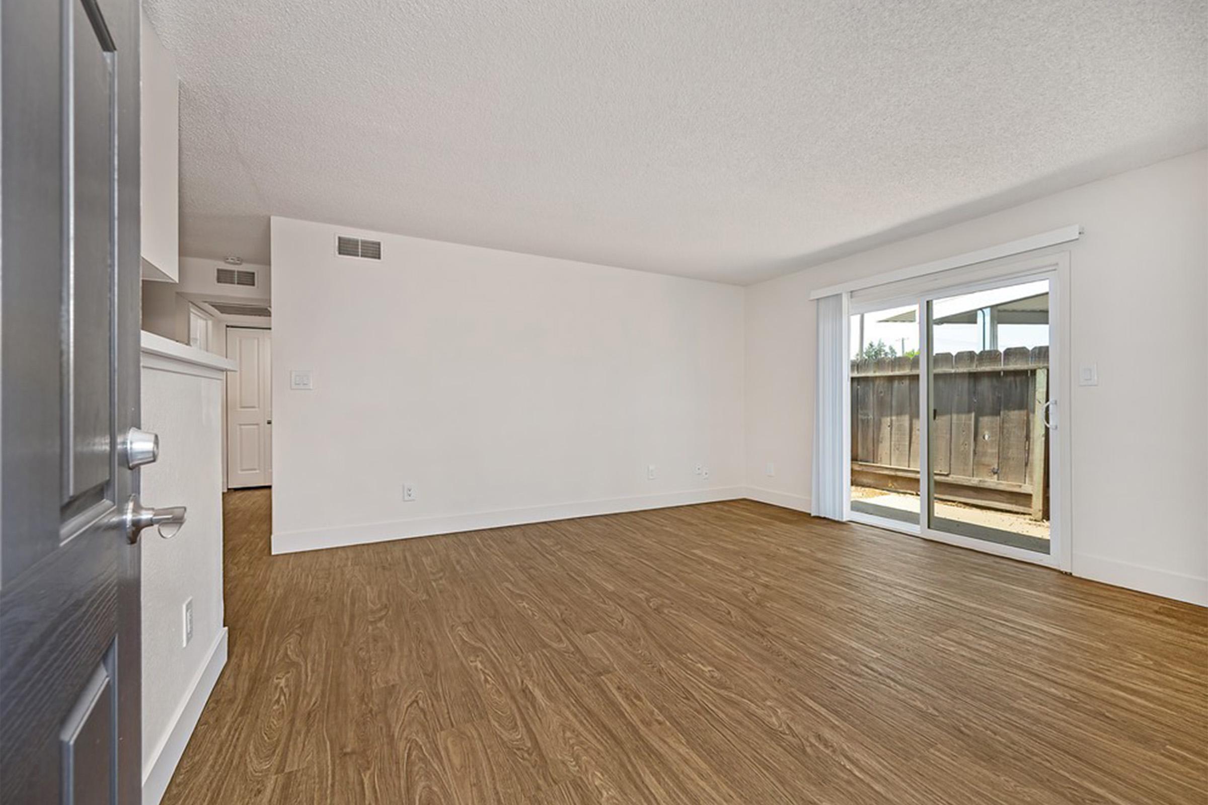 A spacious, empty living room with light wood-style flooring and white walls. A large sliding glass door leads to an outdoor patio area. The room has a bright and airy feel, with natural light streaming in through the door. A doorway on the left leads to another room, adding to the home's layout.