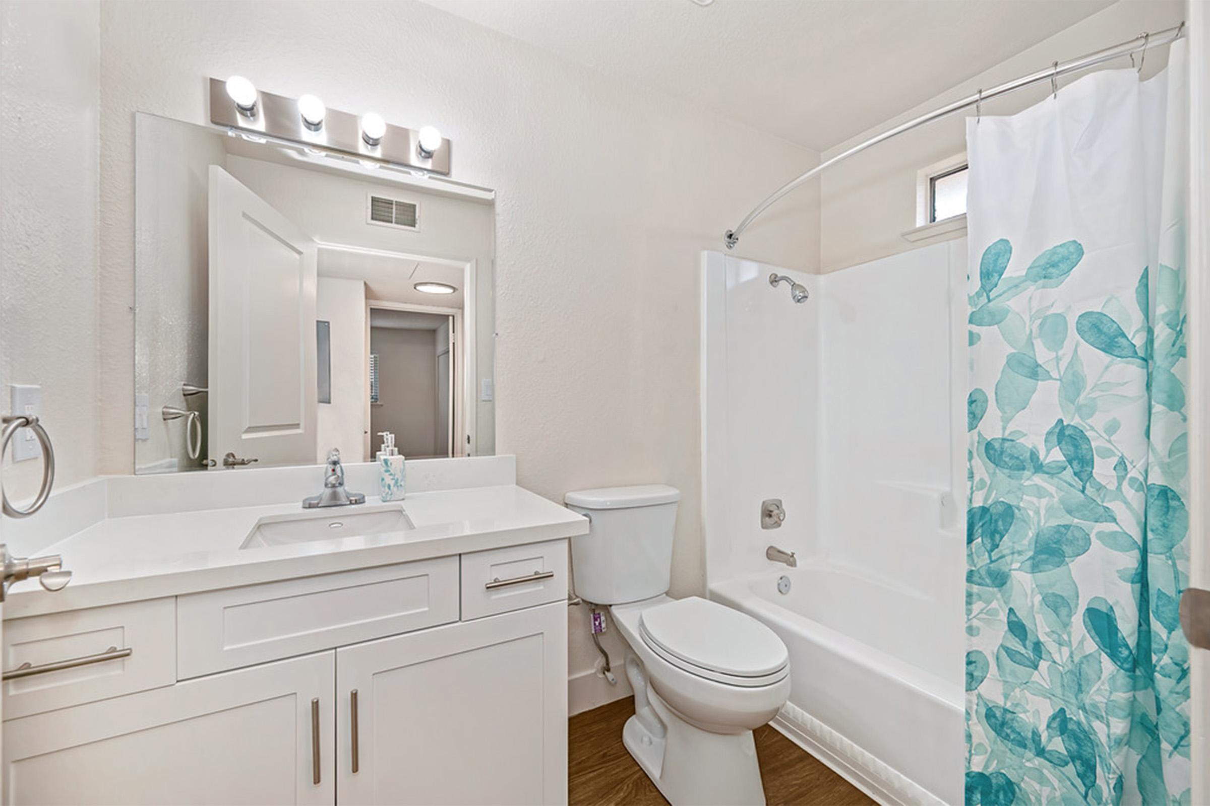 A modern bathroom with a white bathtub, a shower with a clear curtain featuring blue leaf patterns, a white vanity with a sink and a large mirror, and a towel rack. The space is well-lit and has light-colored walls and flooring. A door leading to another room can be seen in the background.