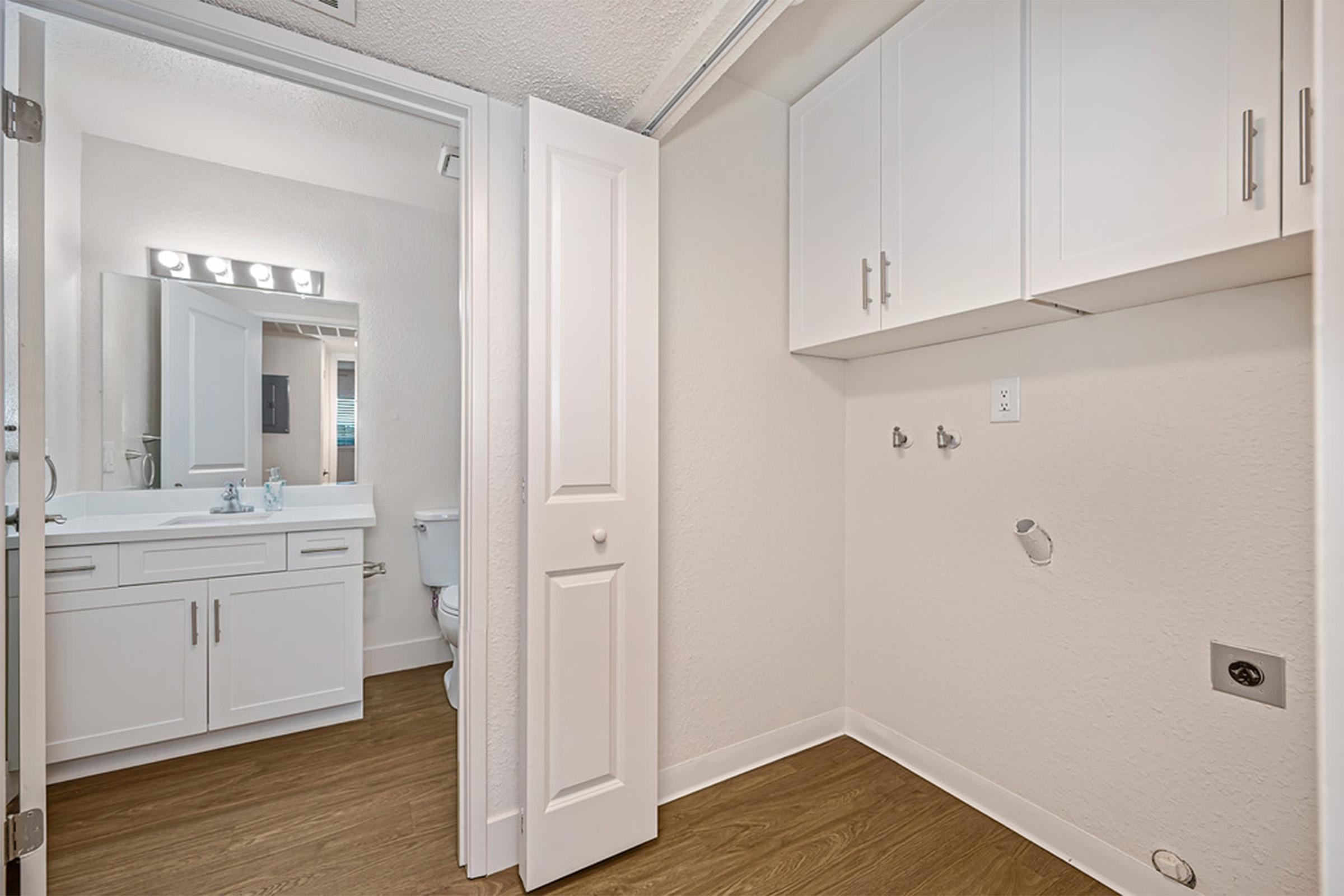 A clean, modern bathroom featuring light-colored walls, a large vanity with a mirror, and a toilet. Adjacent, a small laundry area with a washer/dryer hookup, white cabinets, and wooden flooring. The open door offers a view into the space, enhancing the sense of light and accessibility.