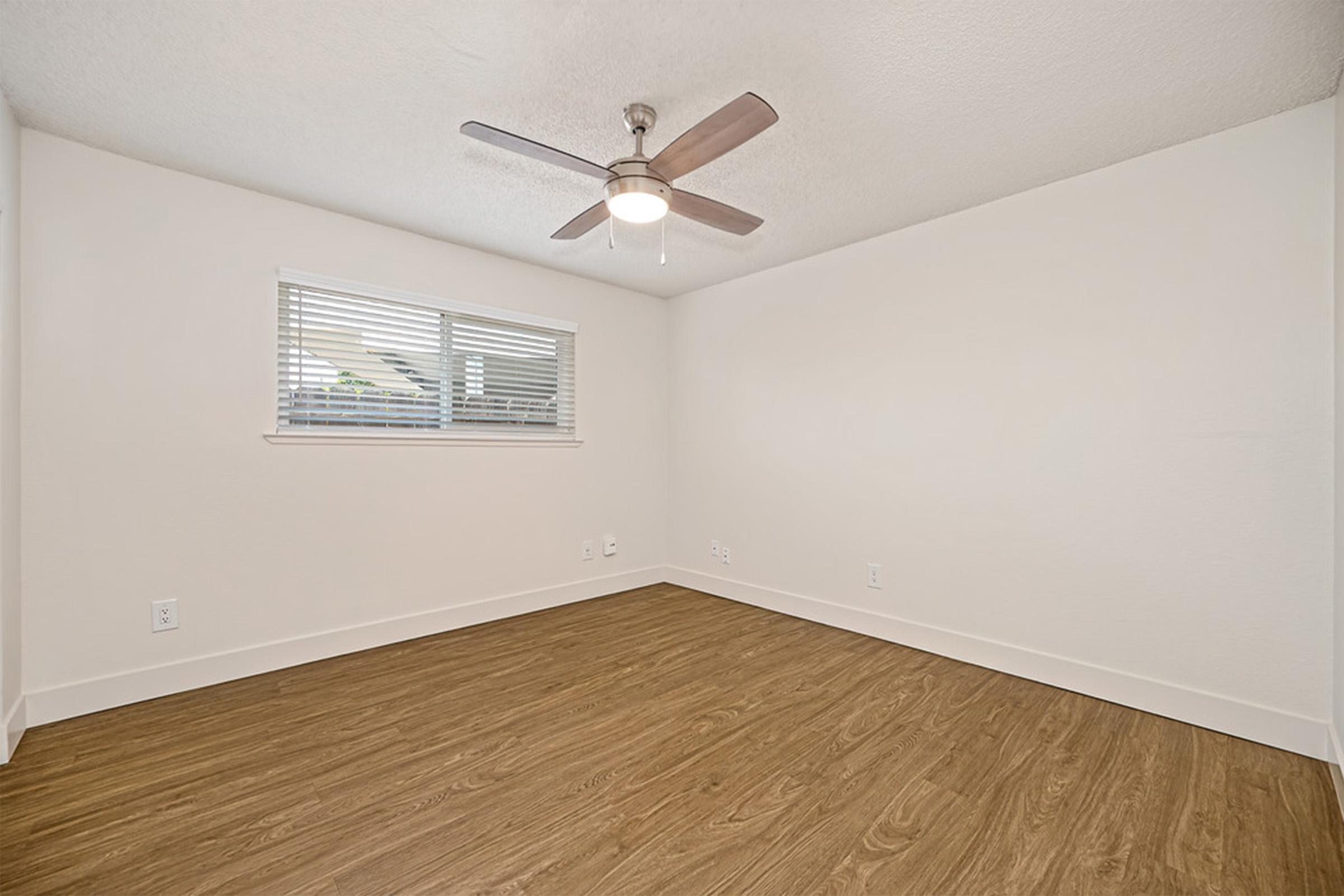 A minimalist empty room featuring light-colored walls and a ceiling fan. There is a window with blinds allowing natural light in, and the floor is covered in wood-like laminate. The overall atmosphere is bright and inviting, suitable for various uses such as a bedroom or office space.
