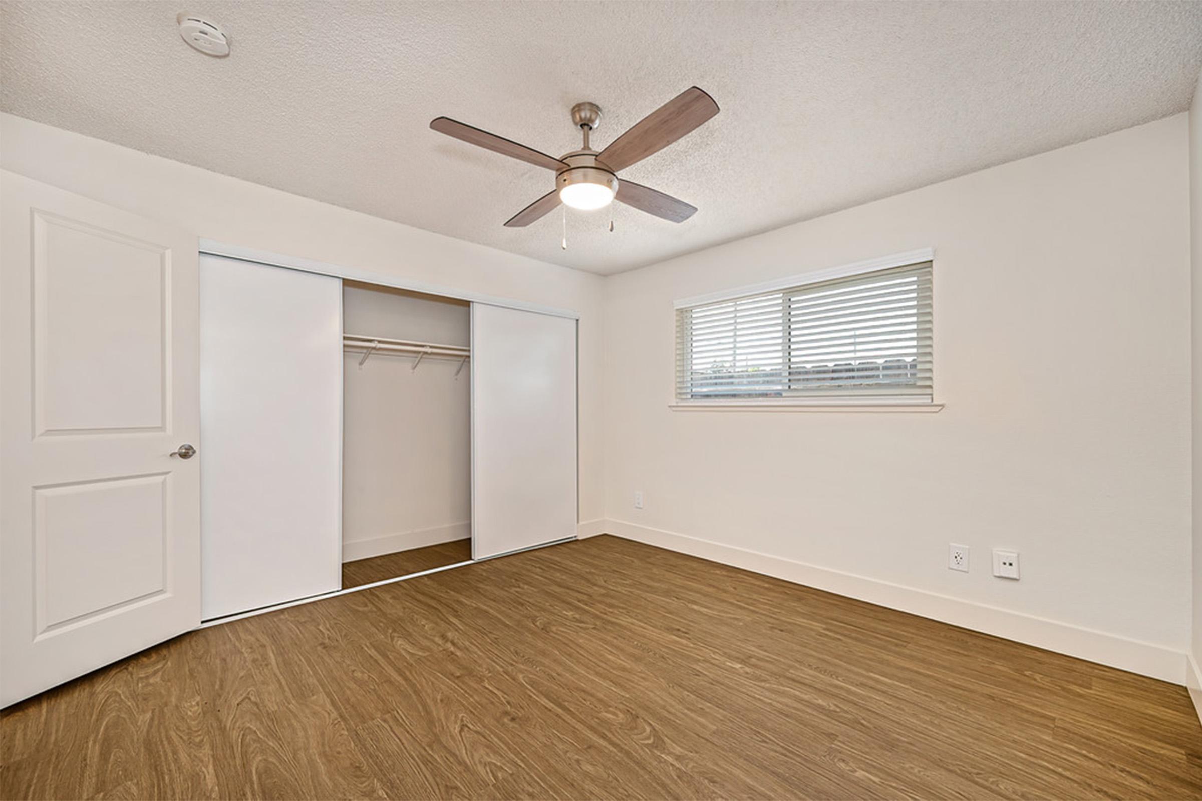 An empty room featuring a ceiling fan, light-colored walls, and a window with blinds. It includes a sliding door closet on one side and a hardwood floor, creating a bright and spacious atmosphere.