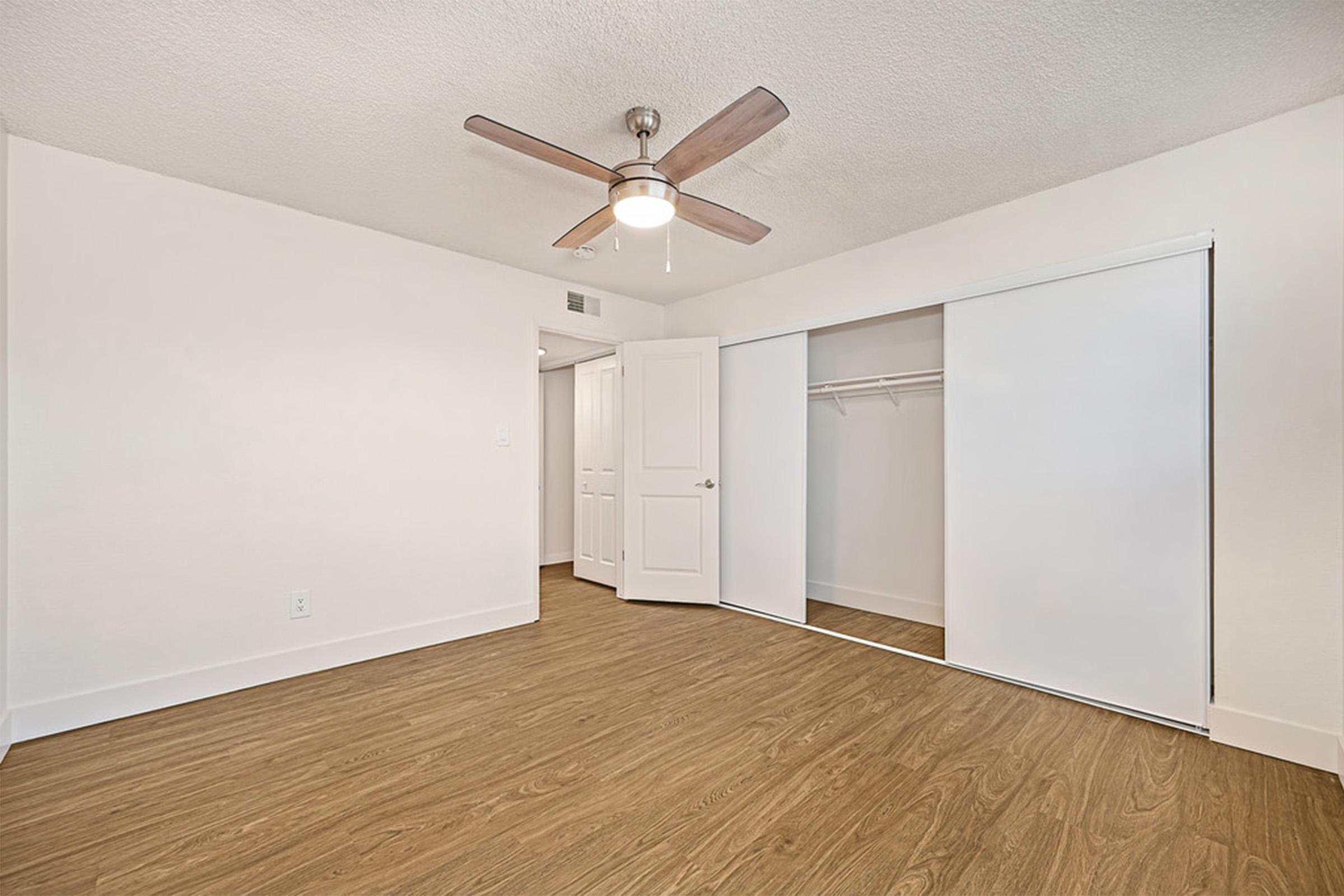 A clean, modern bedroom featuring light-colored walls and a ceiling fan. The room has a sliding closet door on one side, with a mirrored interior. The floor is a warm-toned laminate, and a closed door leads to another space, contributing to a spacious and inviting atmosphere.