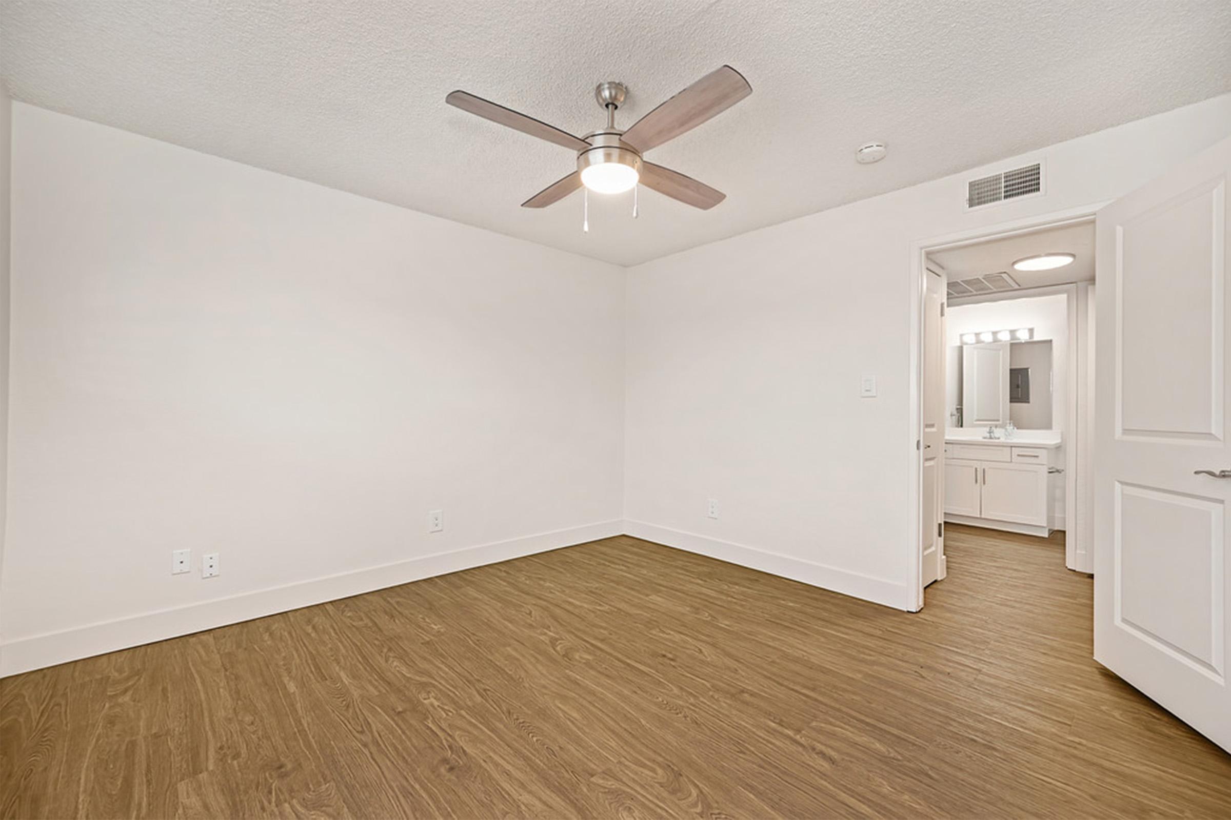 A bright, empty room with light-colored walls and a ceiling fan. The floor is made of wood-like material. To the right, there is an open door leading to a bathroom area, which features a mirror and vanity lighting.