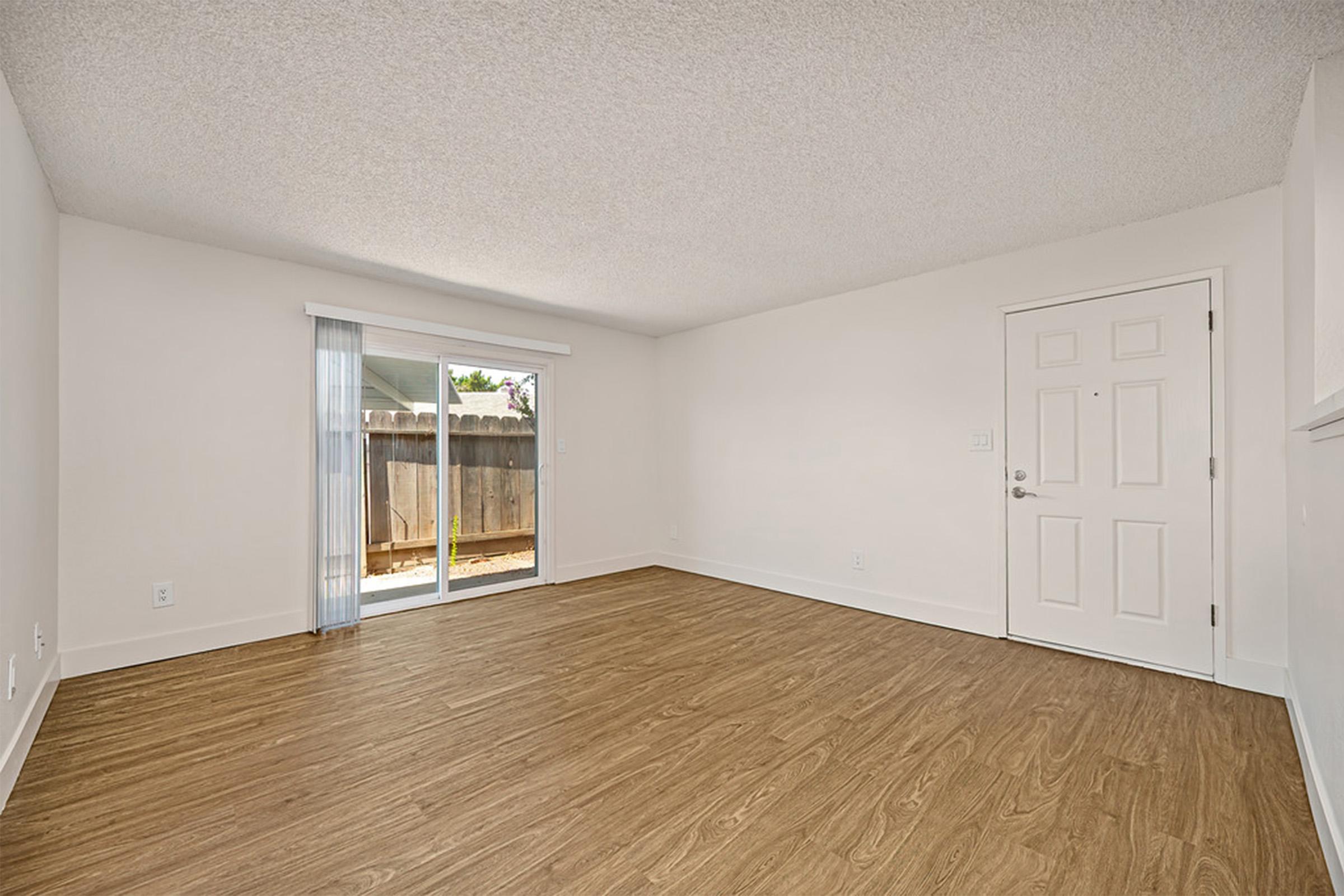 An empty room with a light-colored wall and laminate flooring. A sliding glass door leads to an outdoor area, and there is a simple front door. The space is well-lit with natural light coming through the glass door, creating a neutral and open atmosphere.