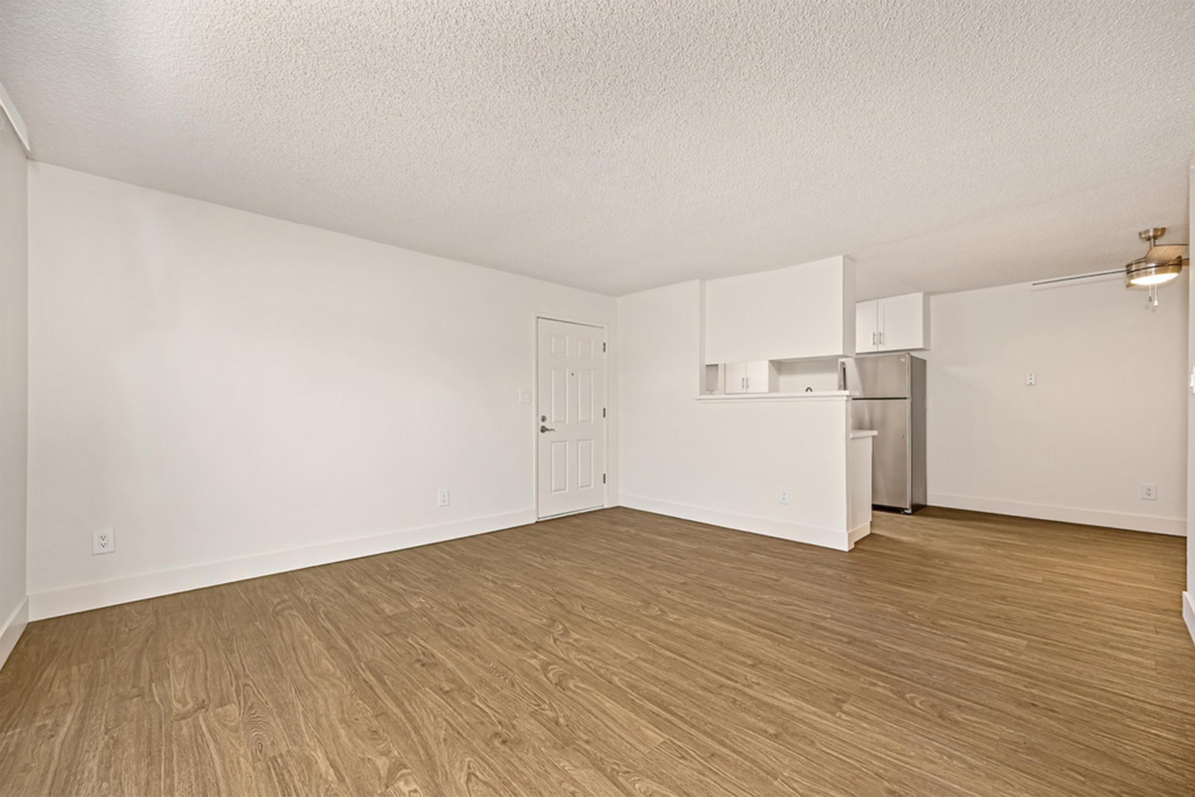 A spacious, empty living area with light-colored walls and wooden flooring. The room features a white kitchen area in the background, including a refrigerator and countertops. Natural light enters through unseen windows, creating a bright atmosphere. The design is modern and minimalistic.