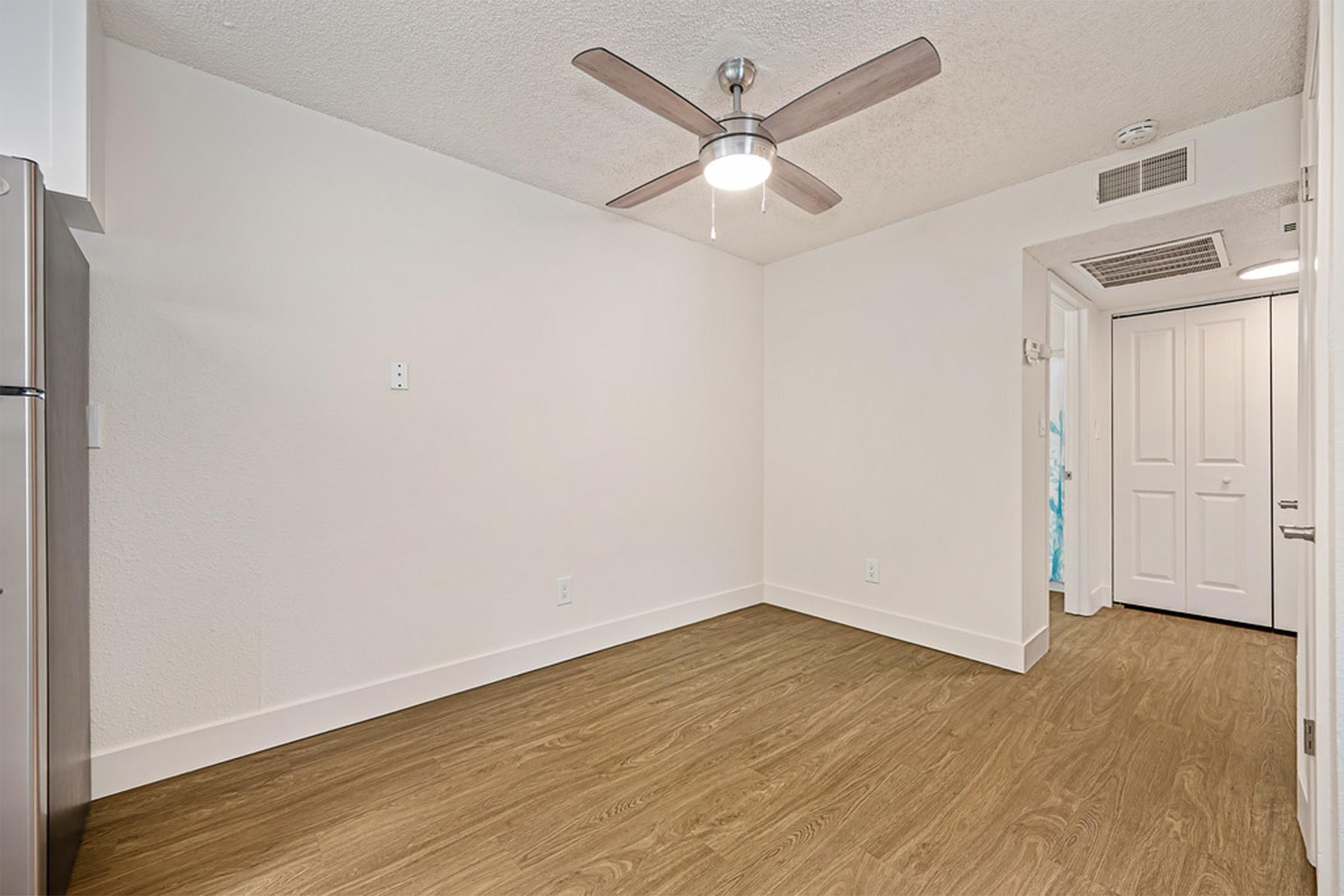 A spacious, well-lit interior of a room featuring a ceiling fan, light wood flooring, and plain white walls. There is a closed door at the far end, leading to another area. The room appears empty and ready for furnishing, conveying a modern and clean aesthetic.
