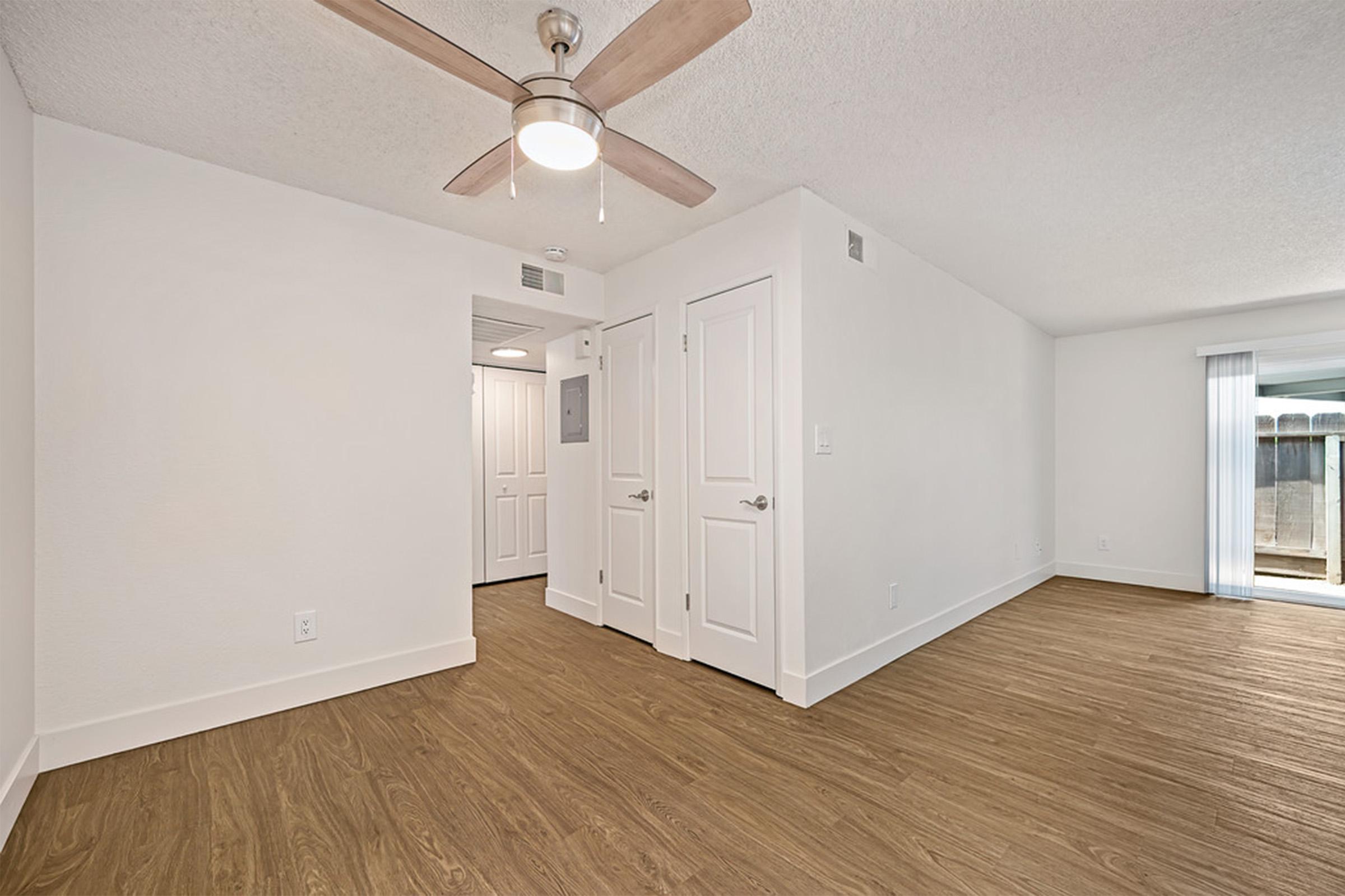 A bright and airy interior space featuring light-colored walls, a ceiling fan, and brown wooden-style flooring. Two white doors lead to other rooms, while a sliding glass door provides access to a patio or outdoor area. The room is well-lit and spacious, creating a welcoming atmosphere.