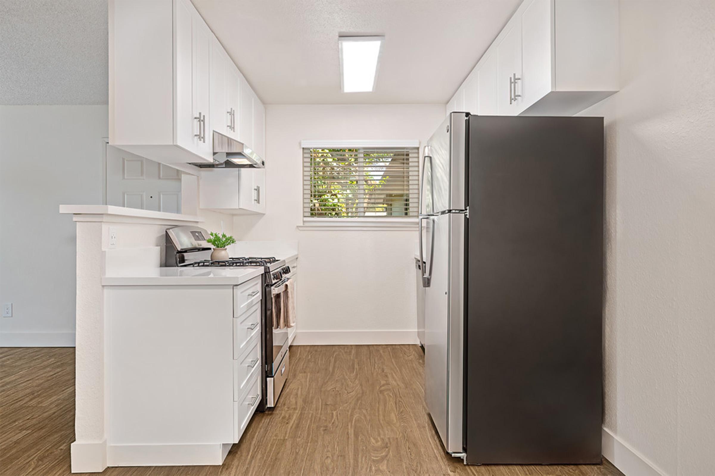 Modern kitchen featuring white cabinetry, a stainless steel refrigerator, and a gas stove. Natural light enters through a window, highlighting the wood-like flooring. The layout is open and clean, creating a bright and inviting atmosphere.