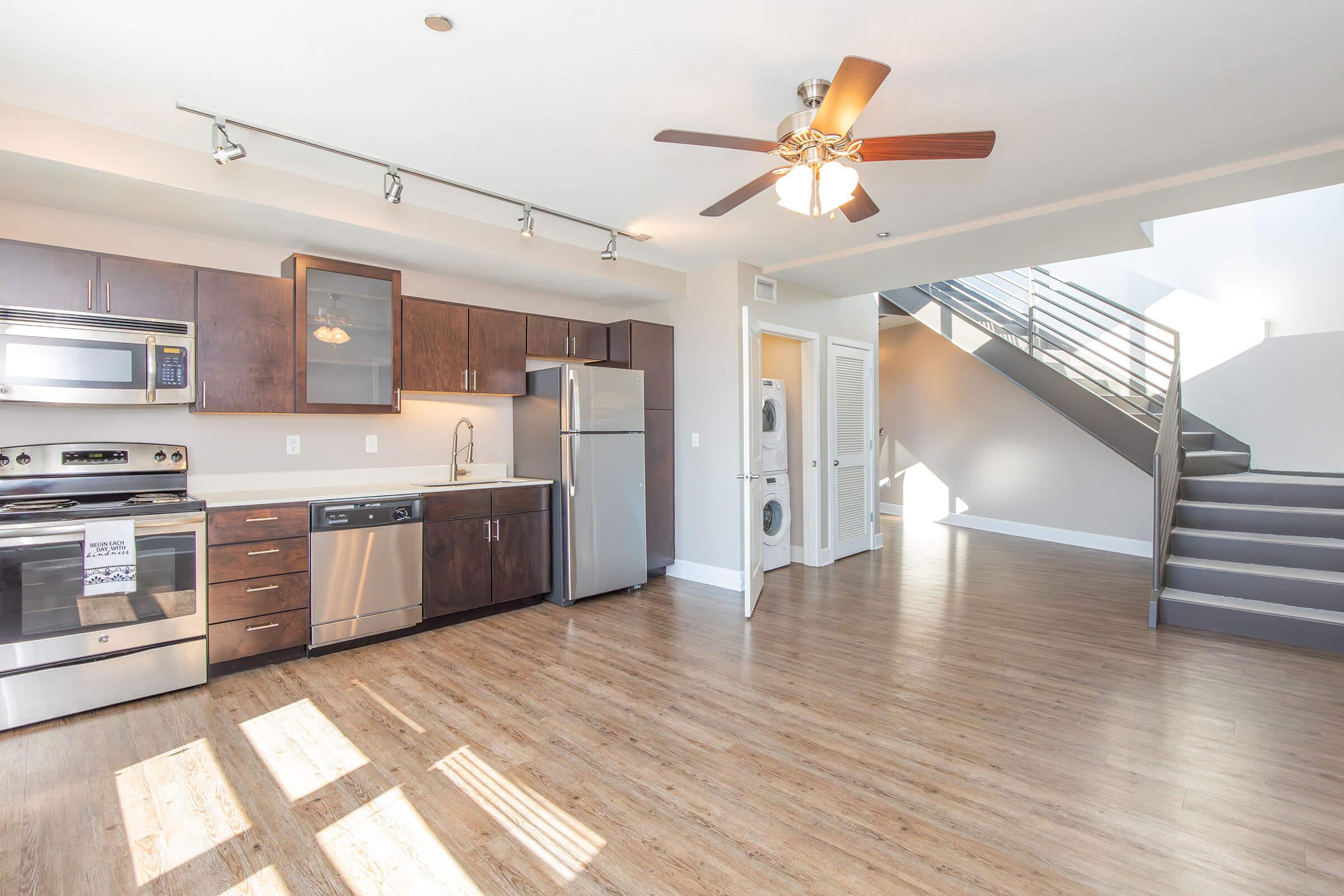 A modern, open-concept kitchen and living area featuring stainless steel appliances, dark wood cabinetry, and a ceiling fan. Natural light floods the space through large windows, highlighting the warm wooden floor. A staircase in the background leads to an upper level, while a laundry area is visibly integrated.