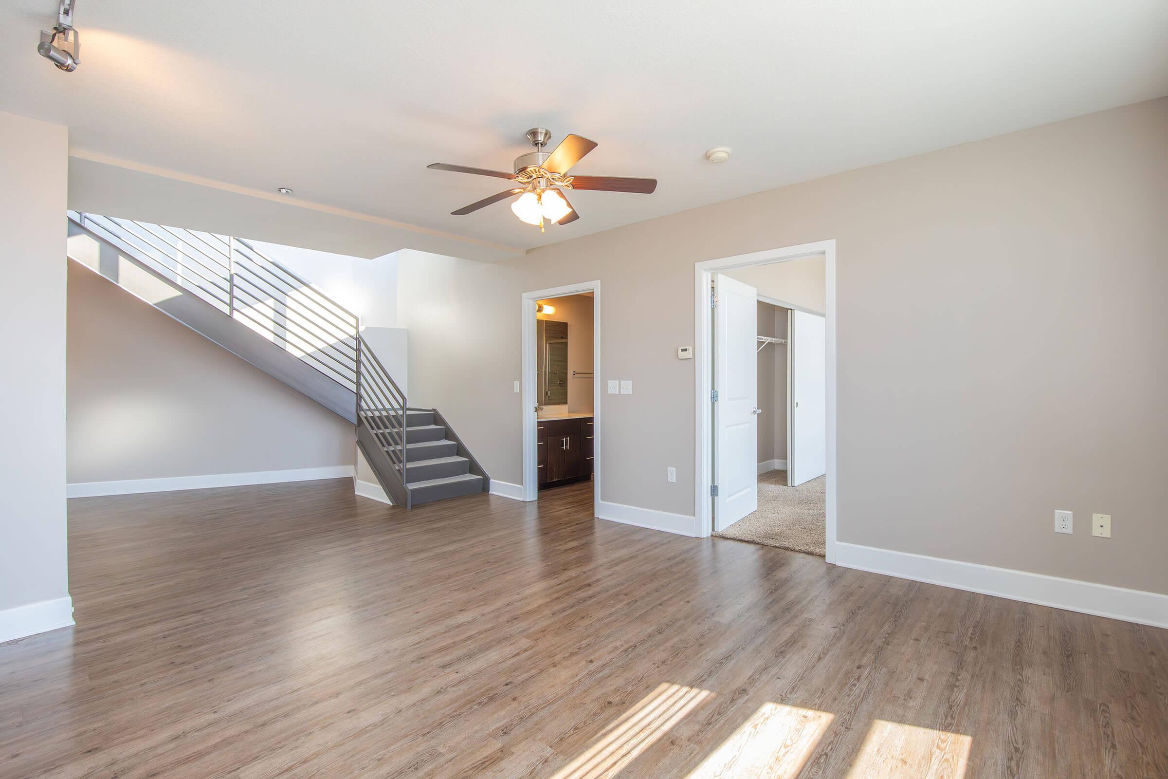 Spacious and modern interior of an apartment featuring a living area with wooden floors, a ceiling fan, and an open layout. A staircase leads to an upper level, while doorways lead to a bathroom and a closet. Natural light fills the space, giving it a bright and inviting atmosphere.