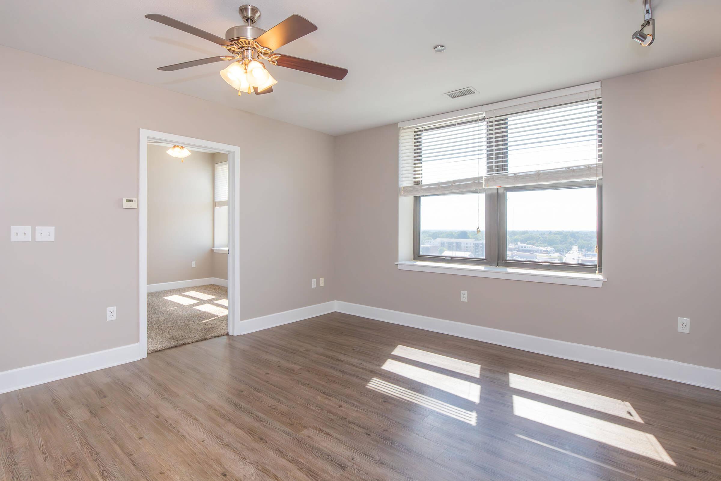 A bright, empty room with light-colored walls and a ceiling fan. Large windows with blinds let in natural light, casting shadows on the wooden floor. A doorway leads to another room in the background, and the space is clean and unfurnished, creating an inviting atmosphere.