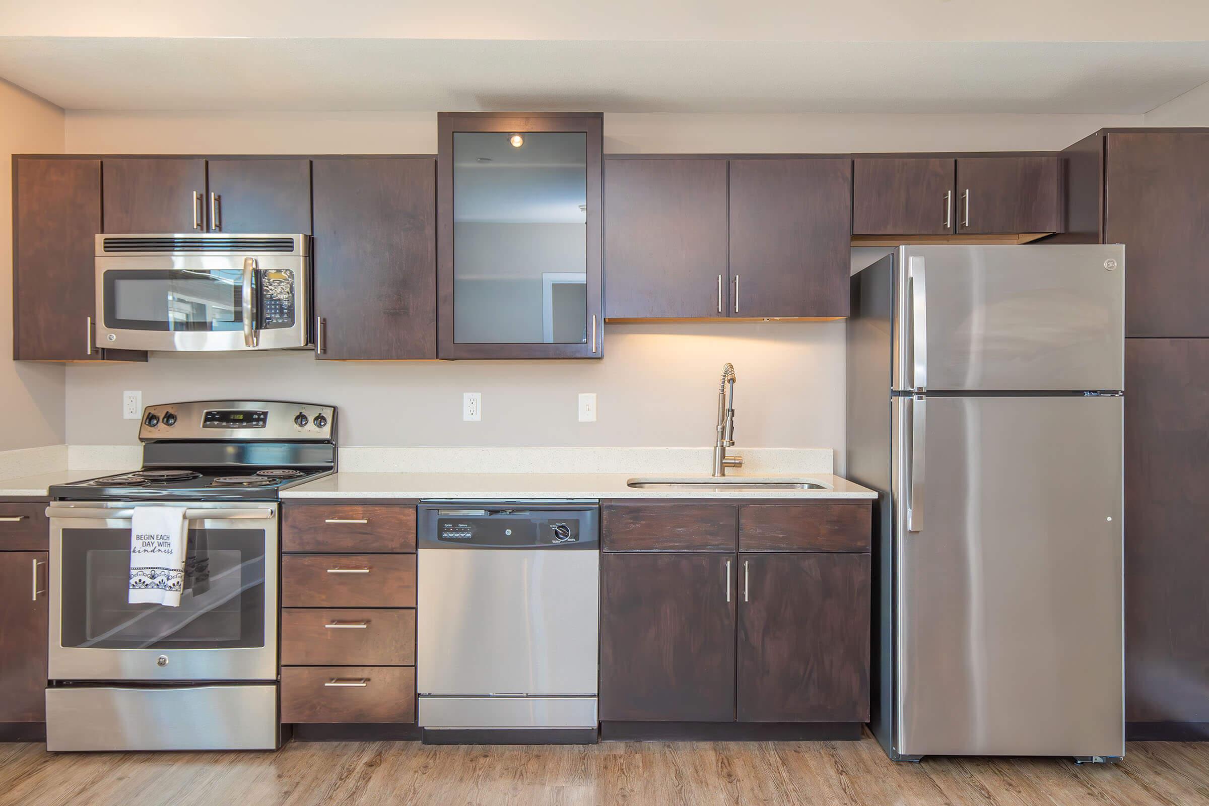 Modern kitchen featuring dark wood cabinets, stainless steel appliances including an oven, microwave, dishwasher, and refrigerator. The countertop is light-colored, and the flooring is wood. The design emphasizes a contemporary and functional space for cooking and meal preparation.