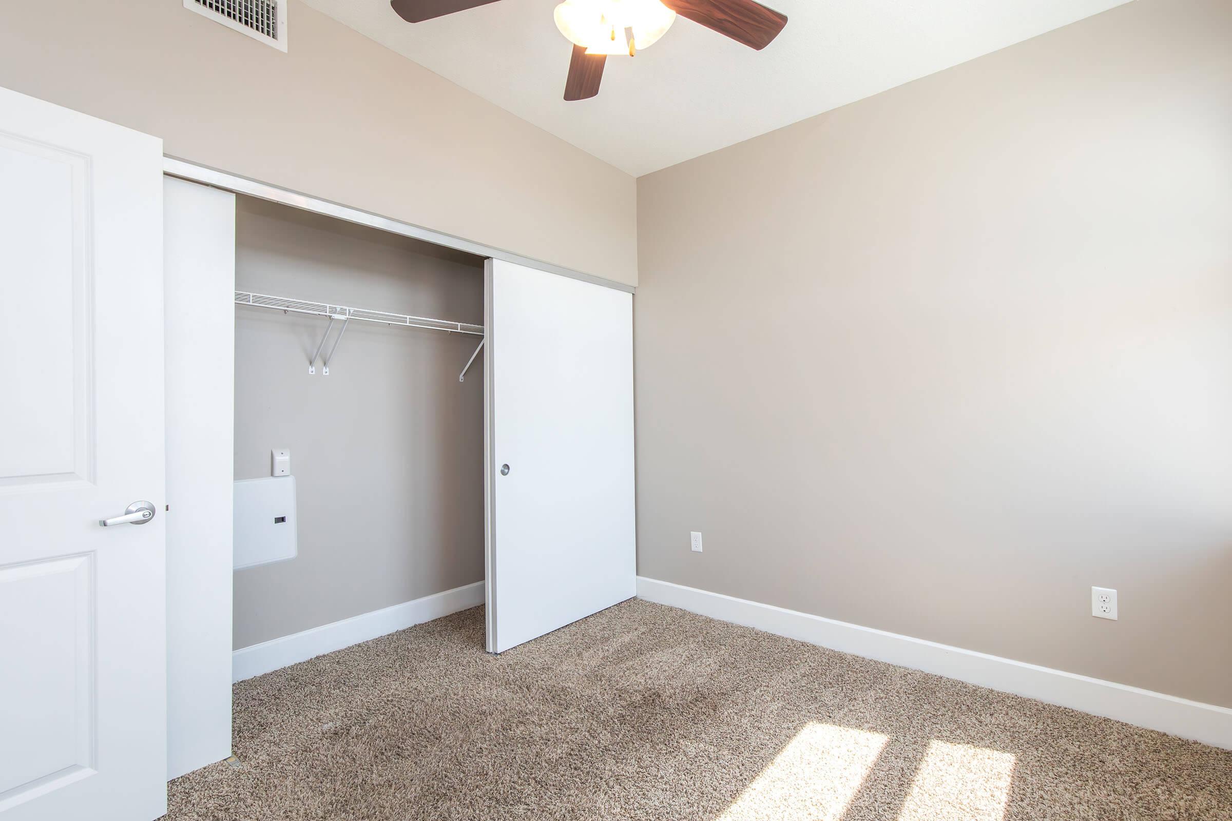 A well-lit, empty bedroom featuring beige walls and carpet. On the left, there is a closet with sliding doors, and a ceiling fan is visible above. Sunlight streams in from a window, creating a warm ambiance in the space.
