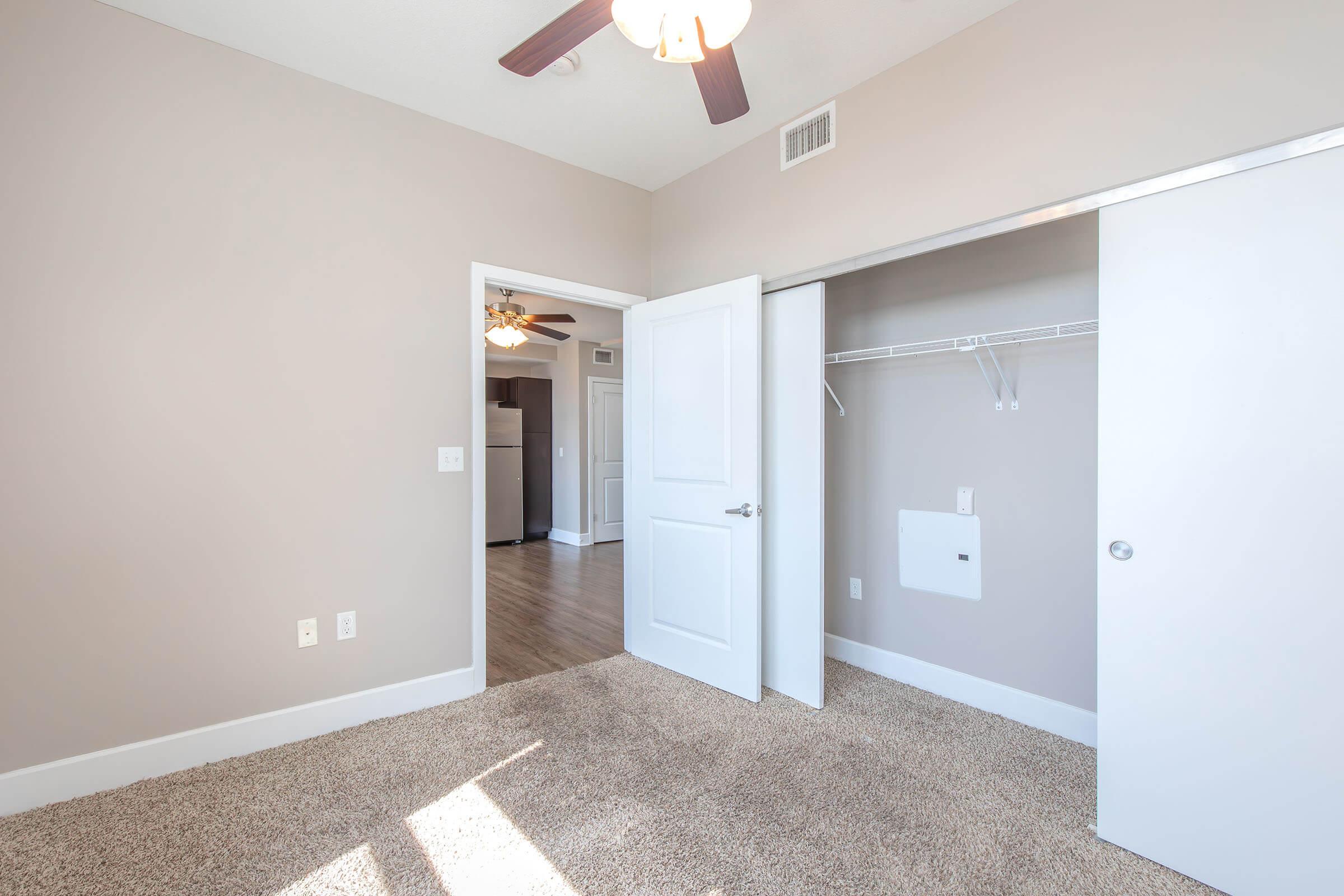A well-lit room with light gray walls and tan carpet. The room features an open door leading to a hallway with a ceiling fan, and a closet with a rod and shelf. Natural light streams in through the doorway, enhancing the spacious feel of the area.