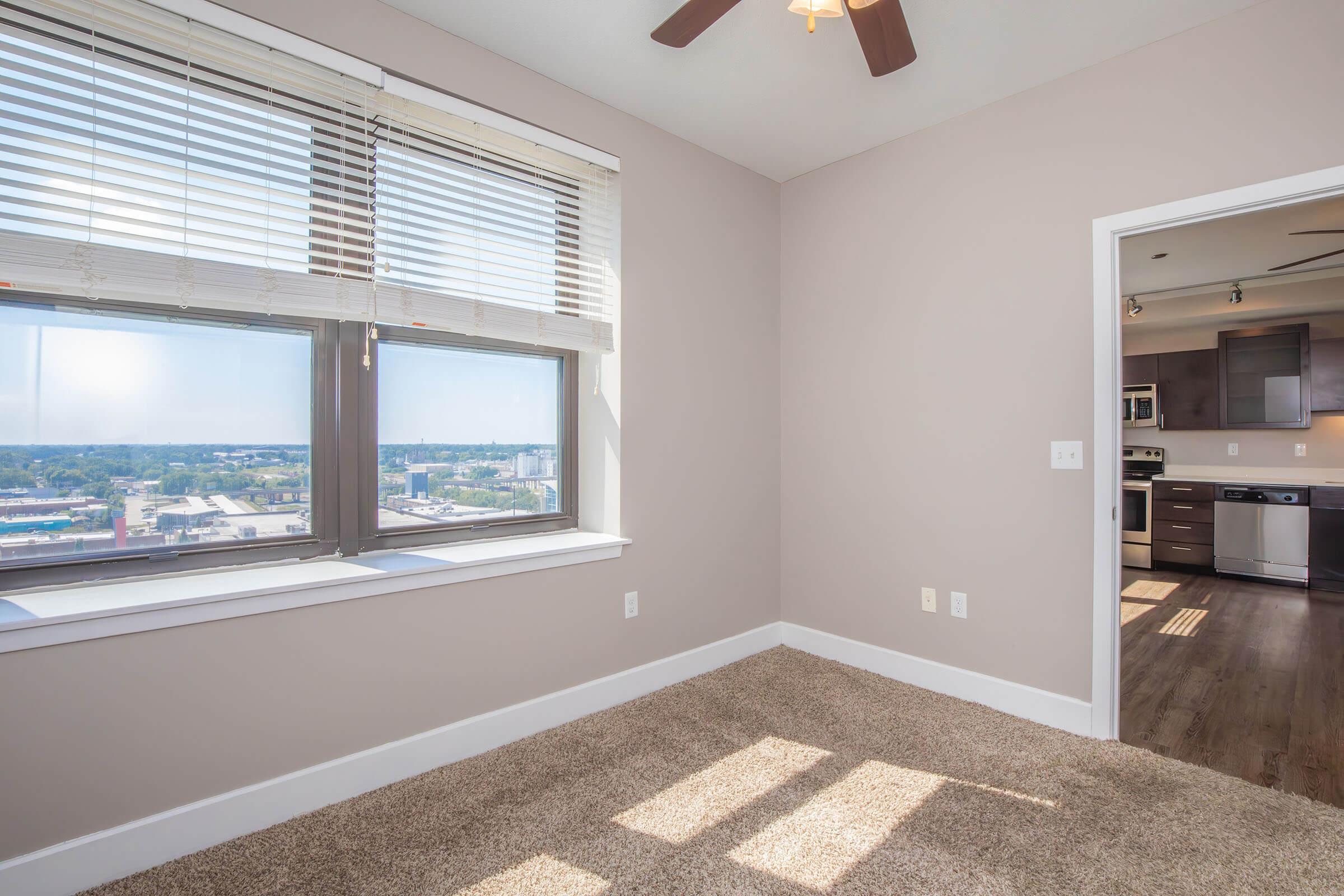 A well-lit, vacant room featuring beige walls and carpeted flooring. It has a large window with blinds providing a view of the city. A ceiling fan is visible. In the background, there is an open doorway leading to a kitchen area with modern appliances. The overall atmosphere is bright and inviting.