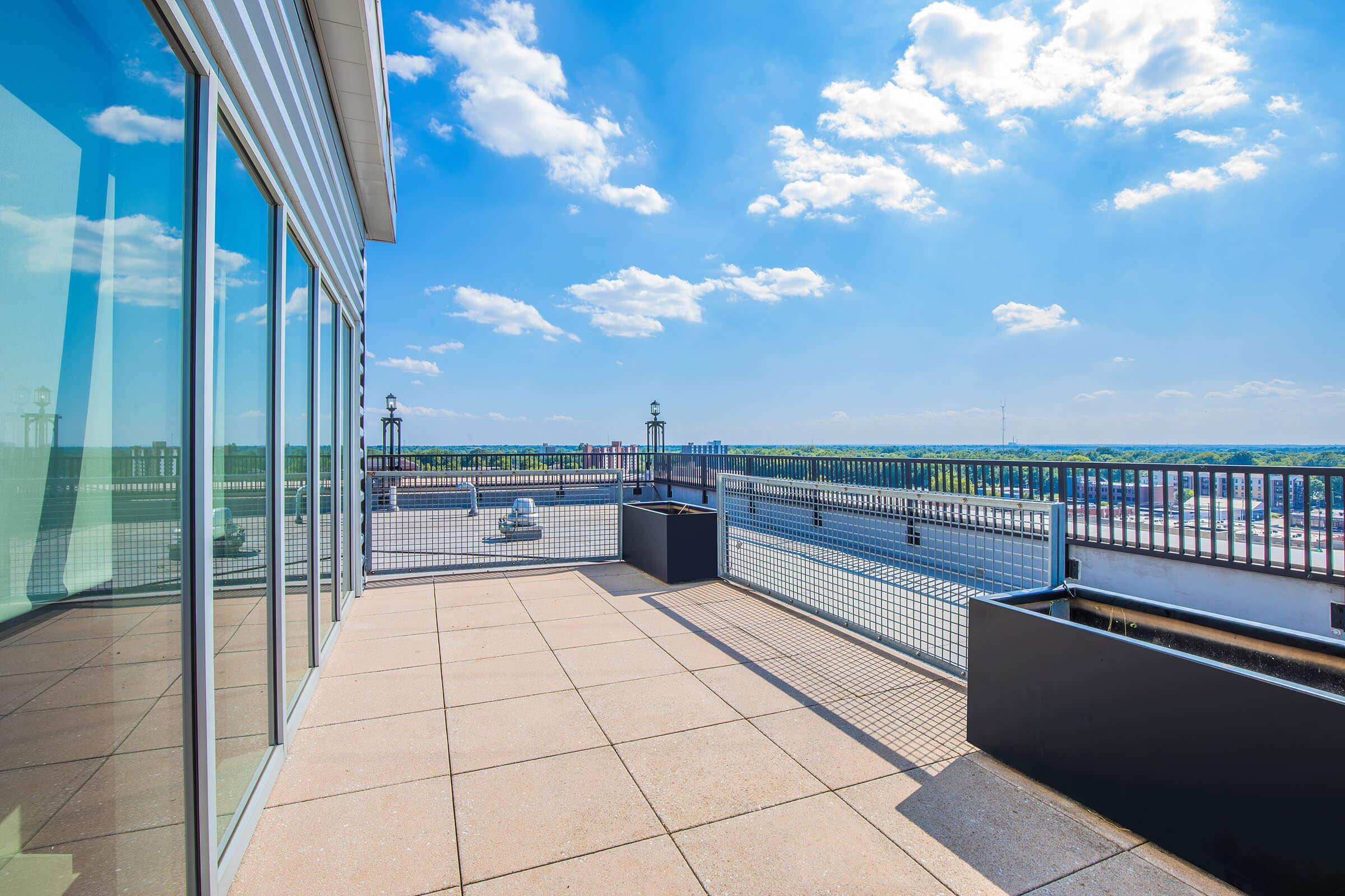 A spacious rooftop terrace with a clear view of the sky and distant landscape. The area is enclosed by a metal railing and features modern flooring. The scene is bright and airy, ideal for relaxation or social gatherings.