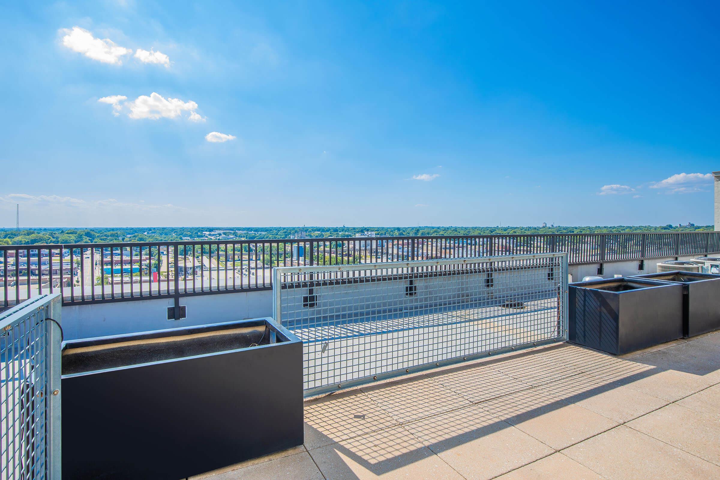 A rooftop view showcasing a clear blue sky with a few clouds, overlooking a landscape of buildings and trees. Black planters line the edge of the rooftop, separated by a metal fence, creating an inviting outdoor space. The scenery highlights a bright and sunny day.