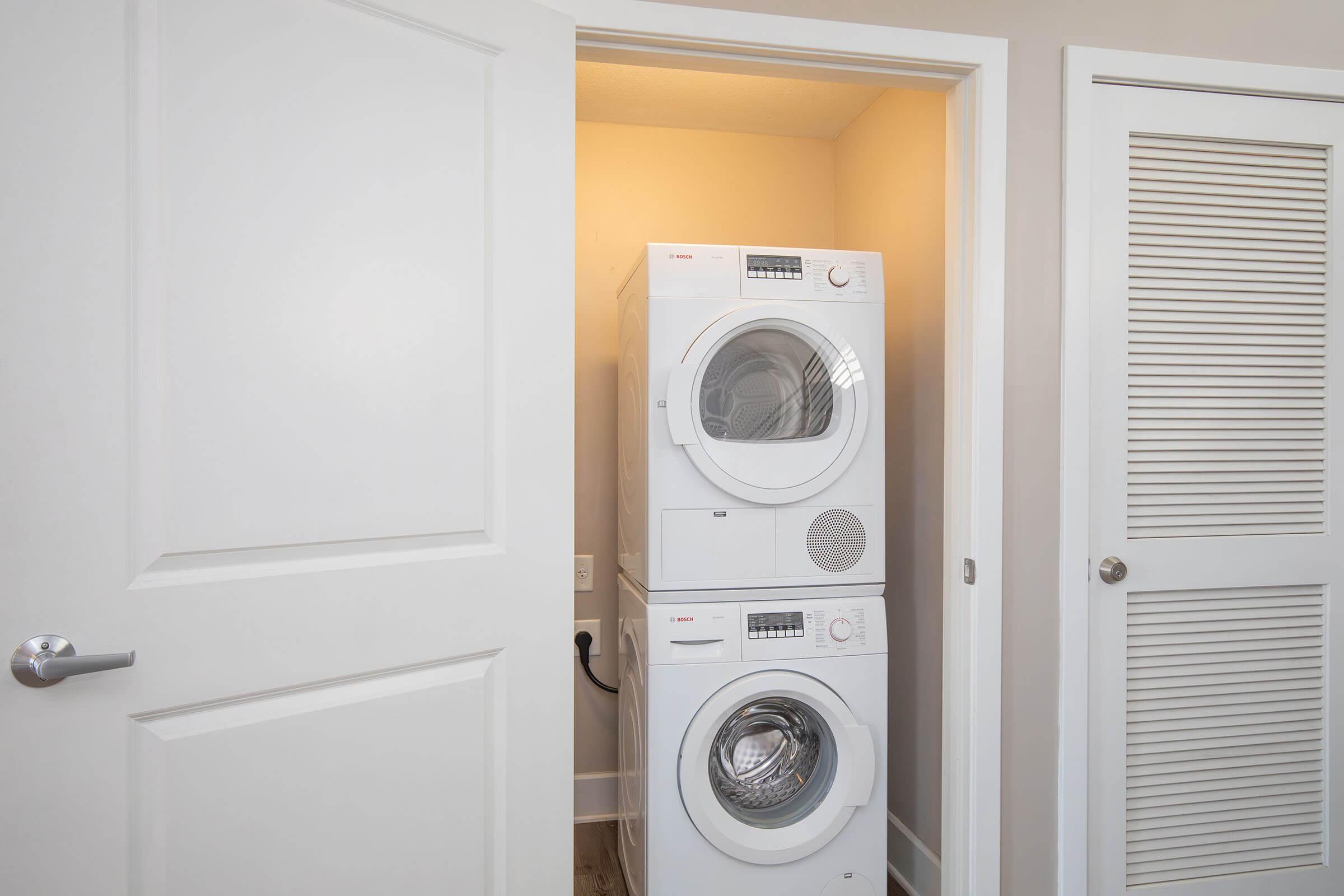 A compact laundry area featuring a stacked washer and dryer unit inside a small room. The doors are partially open, revealing the machines, with light-colored walls and a simple design. One door has a louvered panel for ventilation, while the other is a solid panel.