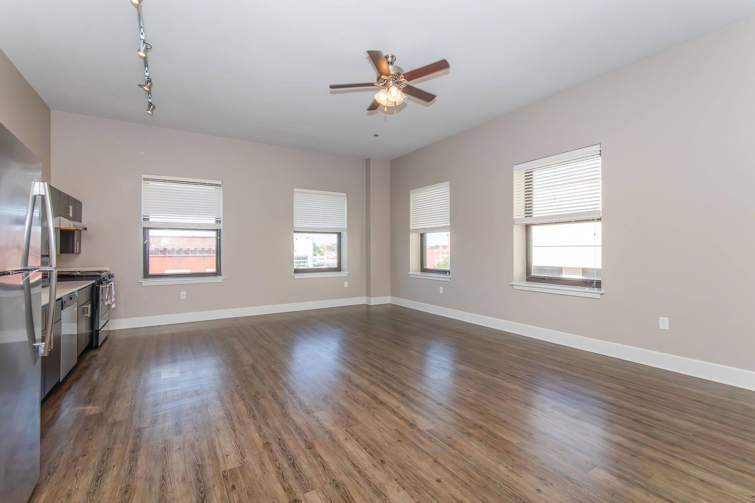 Spacious, unfurnished living area with hardwood floors and large windows. A modern kitchen is visible on the left, featuring stainless steel appliances and a ceiling fan above. Soft beige walls create a warm atmosphere, and ample natural light fills the space.