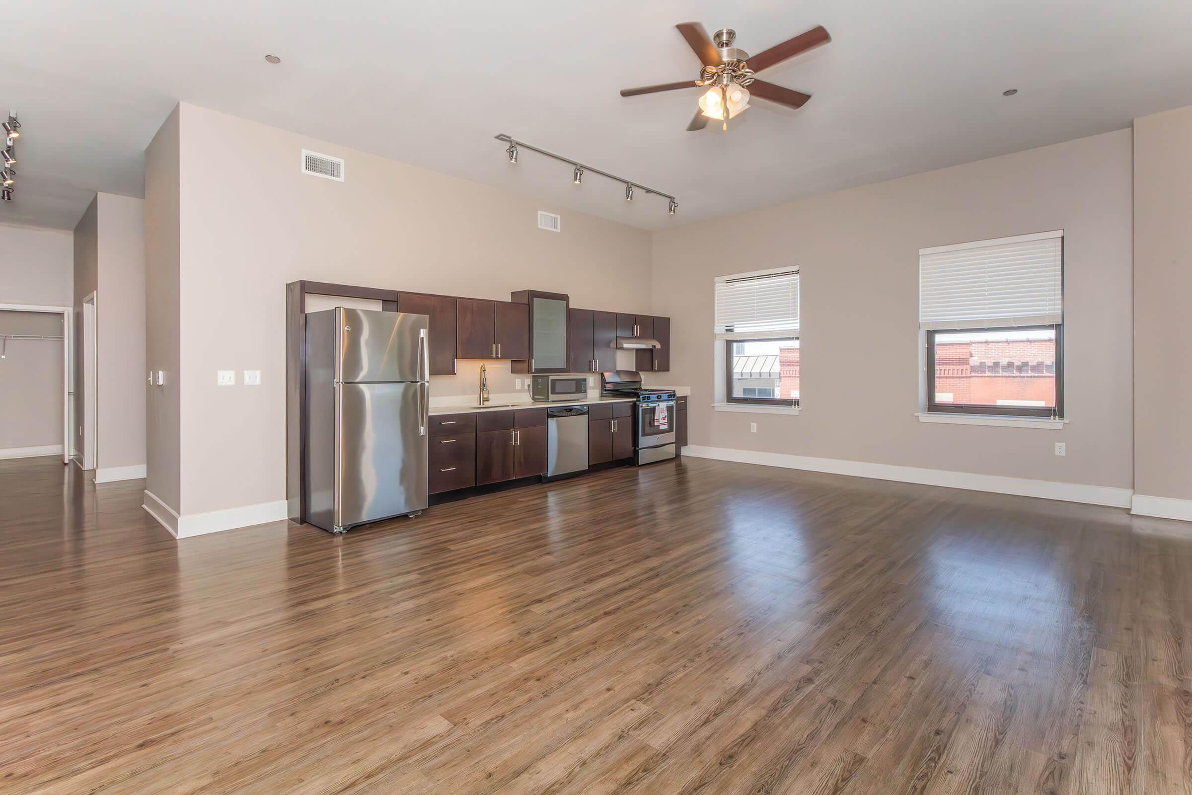 A spacious, modern apartment with hardwood floors, featuring a kitchen area equipped with stainless steel appliances, including a refrigerator, stove, and microwave. The room has large windows allowing natural light, and a ceiling fan. The layout is open with neutral wall colors.