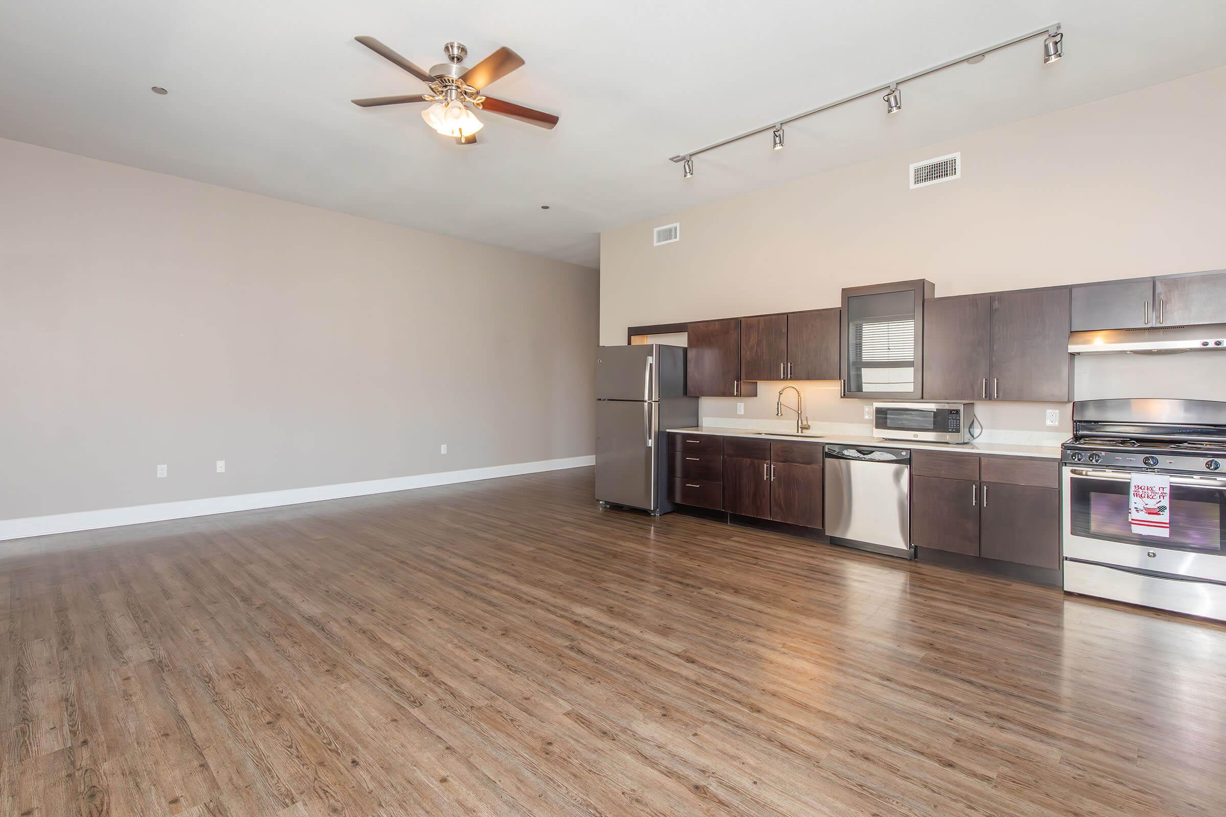Spacious modern kitchen and living area featuring dark wooden cabinets, stainless steel appliances, and a ceiling fan. The room has a light-colored wall and a hardwood floor, creating a bright and open atmosphere. Large windows allow natural light to fill the space.