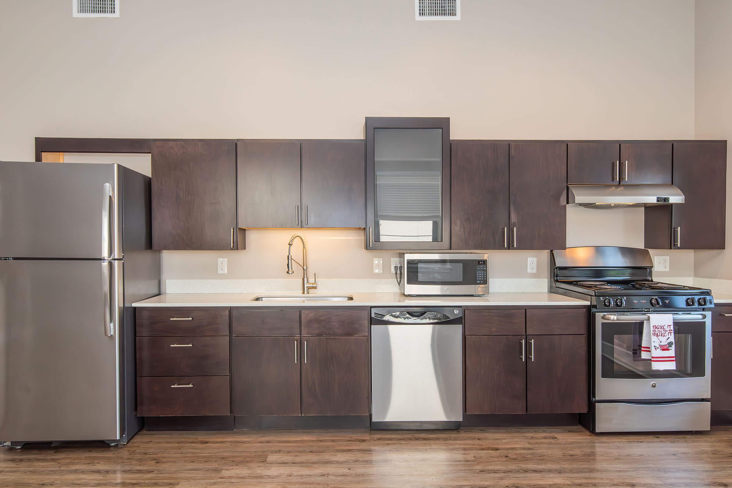 A modern kitchen featuring dark wood cabinets, stainless steel appliances including a refrigerator, microwave, and gas stove. The countertop is light-colored and there is a sink with a sleek faucet. The floor is wooden, and the overall design is contemporary with an open and spacious feel.