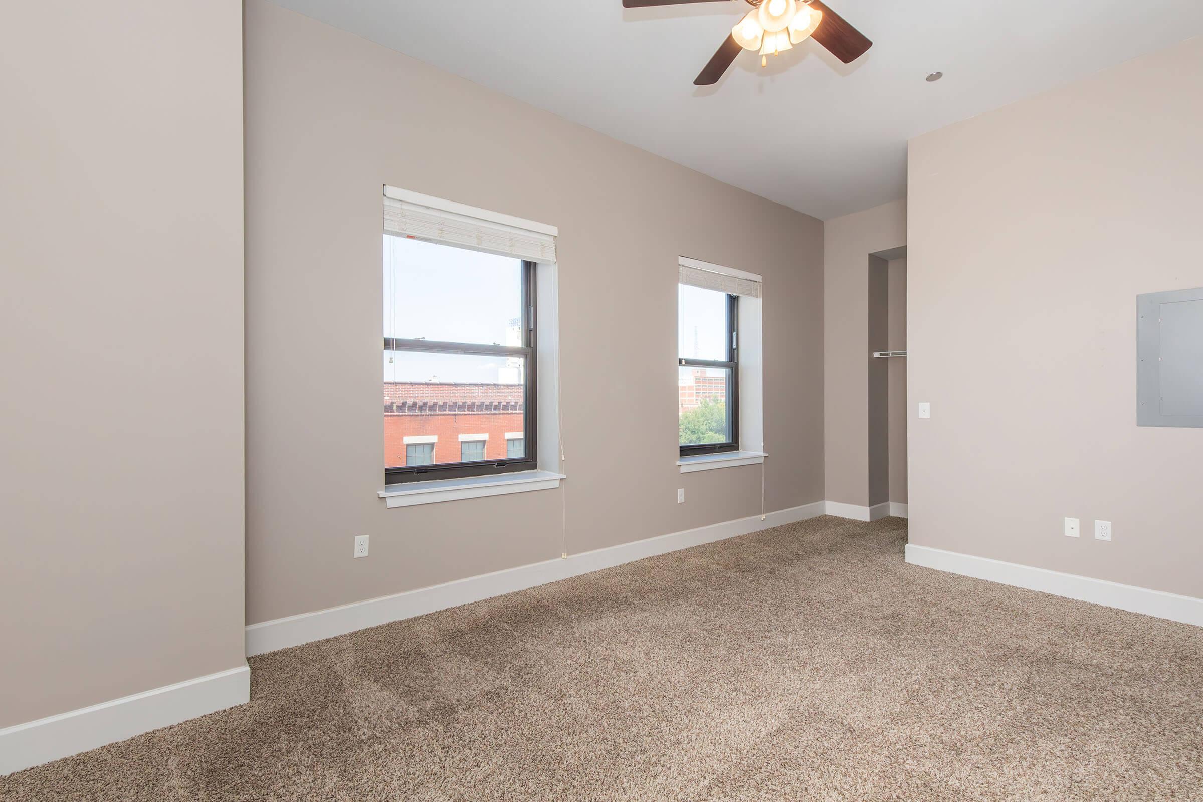 A spacious, empty room with light beige walls and brown carpet. It features two windows with white blinds allowing natural light, a ceiling fan, and a closet space visible to the right. The room has a modern, minimalist feel, without any furniture or decorations.