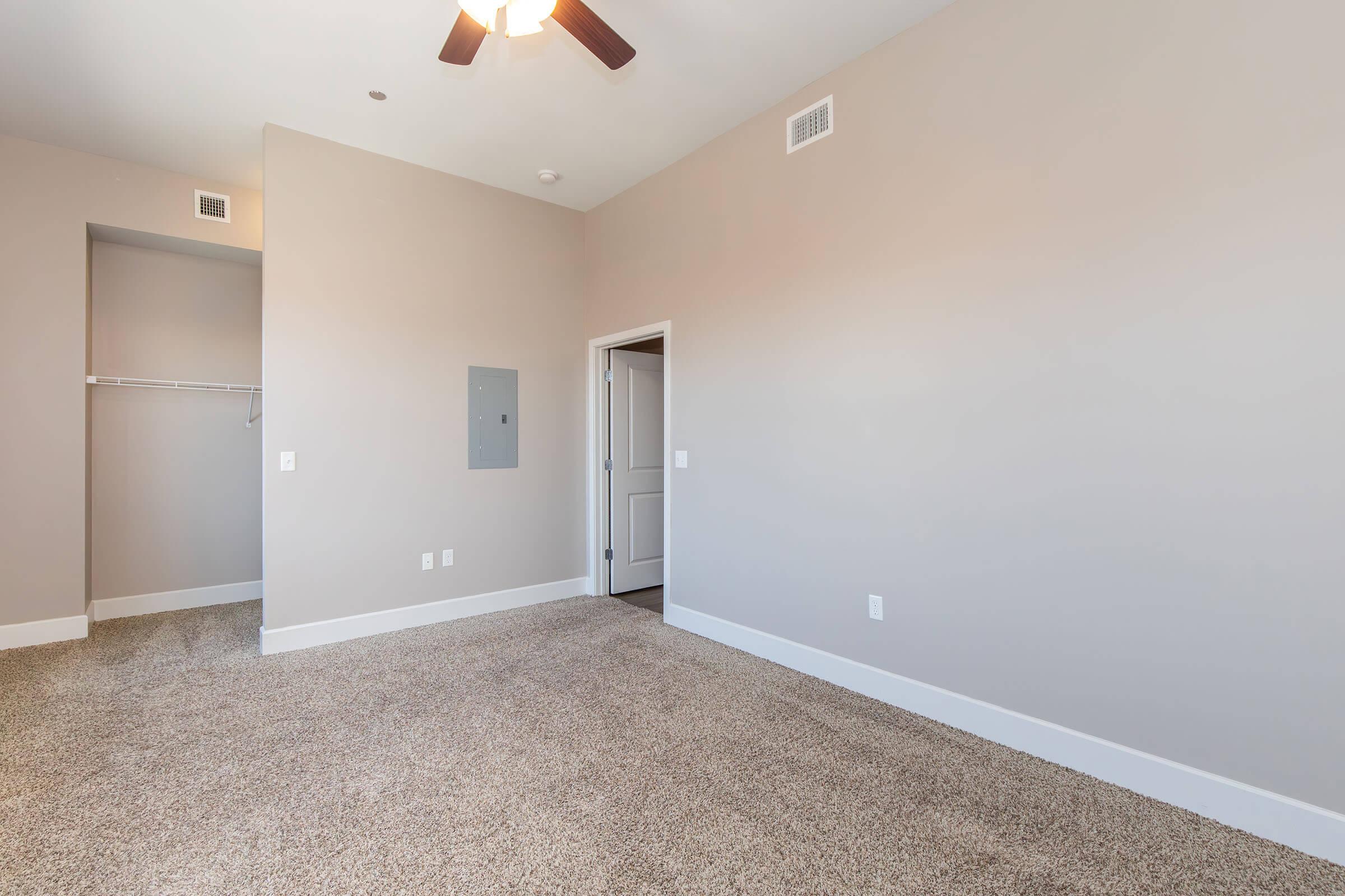 A spacious, light-colored room with beige walls and a ceiling fan. Carpeted flooring, a small closet area on the left, and a door leading to another space visible. The room is empty, creating a bright and open atmosphere.