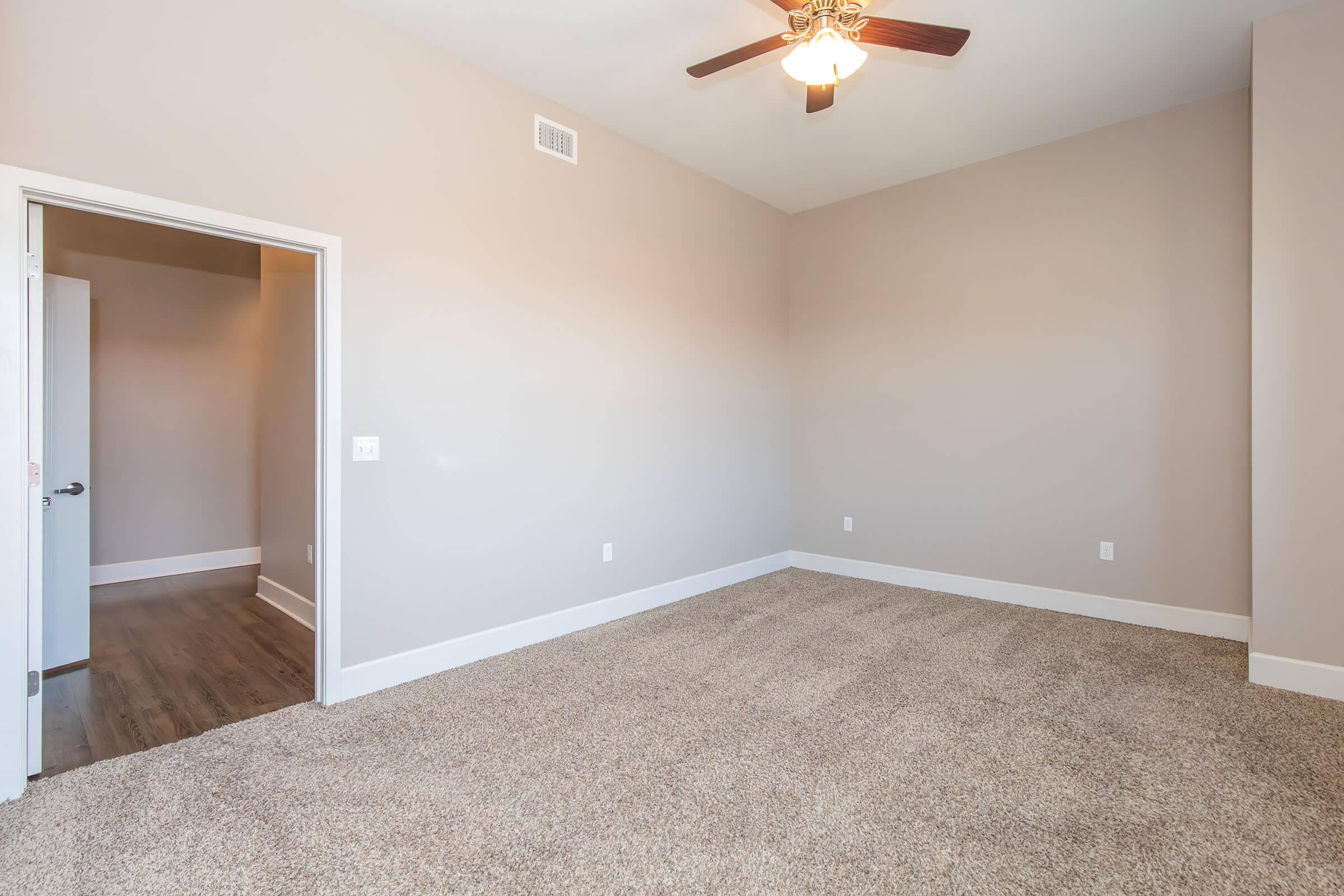 A spacious and well-lit empty room with beige walls and carpeting. It features a ceiling fan and a doorway leading to another room. The floor is hardwood, and the design is modern and minimalist, creating an inviting atmosphere.