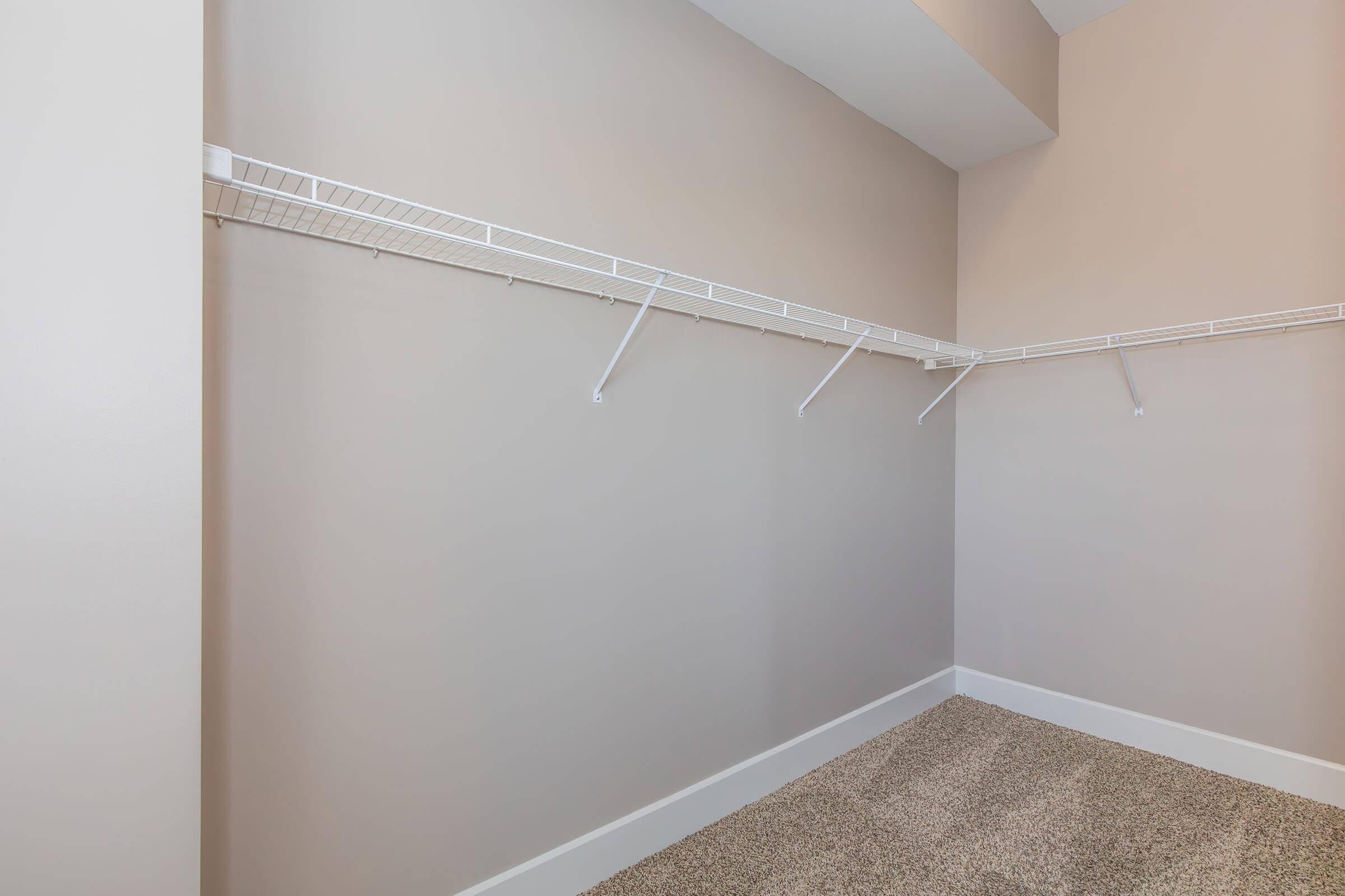 Empty closet with beige walls and a light brown carpet. The closet features wire shelving along one wall, with several brackets but no items stored. The overall space is well-lit and clean, making it ideal for organization or storage.