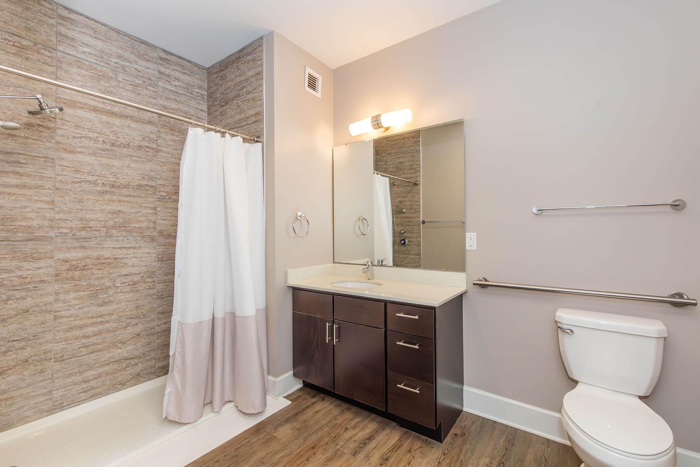 A modern bathroom featuring a walk-in shower with a white curtain, a beige countertop sink with dark wood cabinetry, a large mirror, and a toilet. The walls have a neutral color, and the floor is covered in light wood-like tiles. There are grab bars for safety near the toilet and shower area.