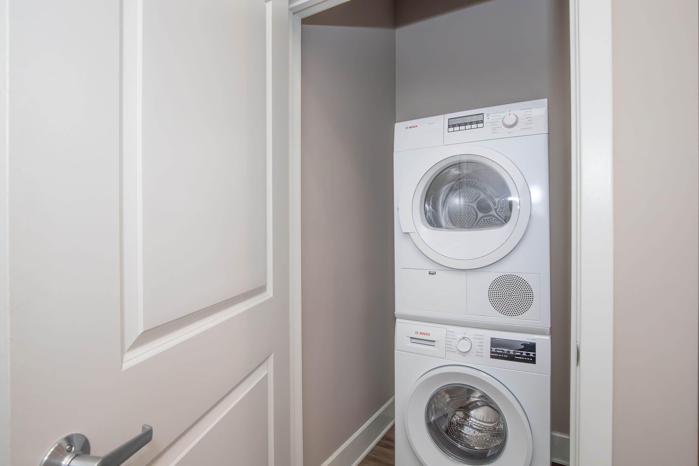 A stacked washer and dryer set located in a small closet space. The washer is on the bottom and the dryer is on top. The closet has a light-colored wall and a door that is slightly open, revealing the appliances inside. The flooring appears to be wooden.
