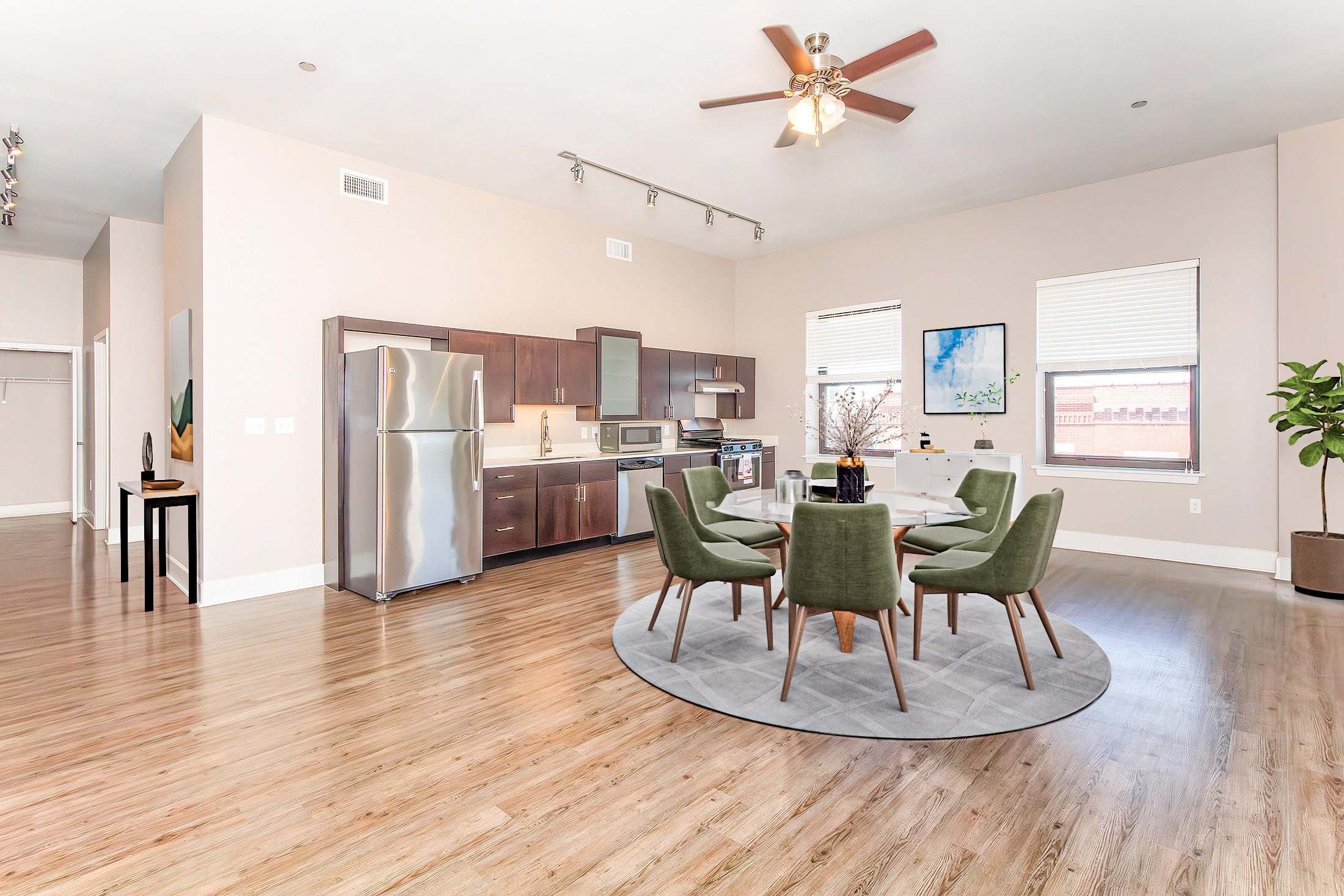 Modern open-concept kitchen and dining area featuring hardwood floors, stainless steel appliances, and a circular gray area rug. A round dining table is surrounded by four green chairs, with large windows providing natural light. The space is decorated with minimalistic art and a potted plant.