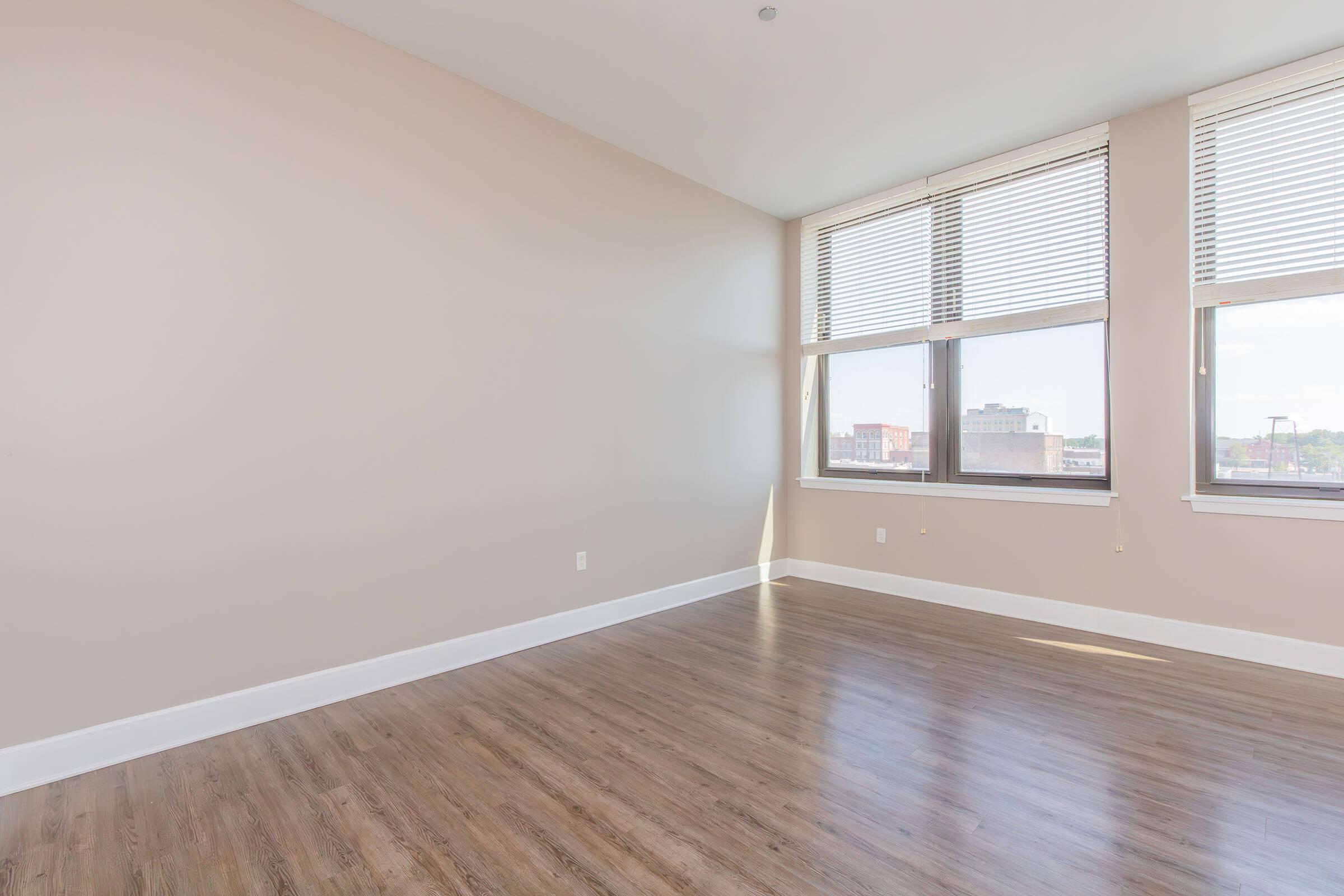 A spacious, empty room with light beige walls and large windows adorned with white blinds. The floor is made of wooden planks, and the overall atmosphere is bright and airy, receiving ample natural light from the windows. The room has a minimalist design, suitable for various uses.