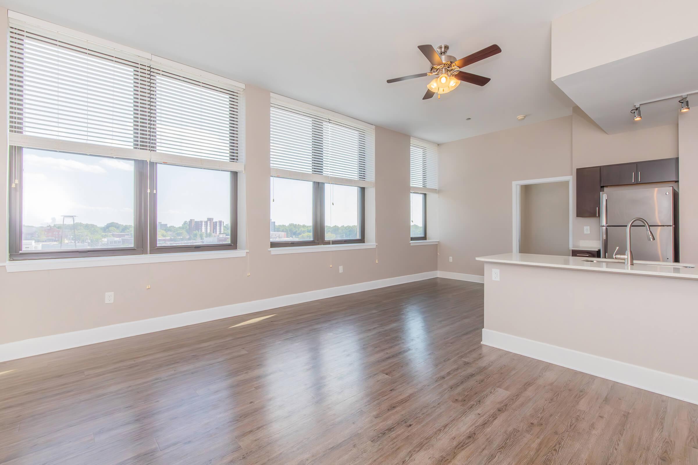 Spacious modern apartment interior with large windows, hardwood flooring, and a ceiling fan. Features an open layout with a kitchen area visible in the background, showcasing stainless steel appliances and a bright, inviting atmosphere.
