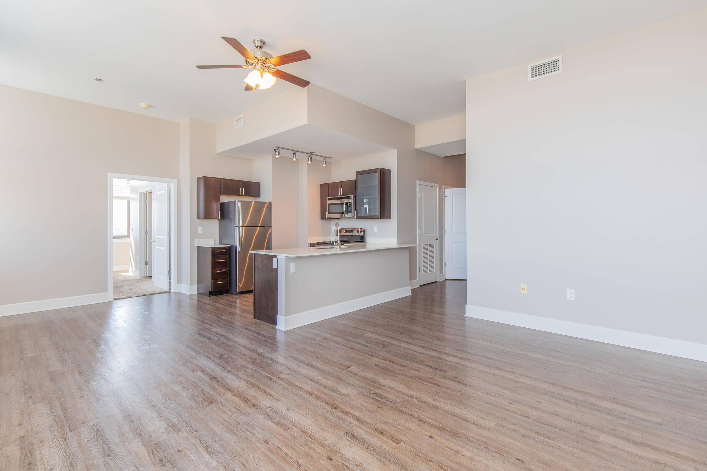 A spacious modern apartment featuring a hardwood floor, ceiling fan, and an open kitchen. The kitchen has dark cabinets, stainless steel appliances, and a breakfast bar. There are bright walls and natural light from a nearby window. Doorways lead to additional rooms, enhancing the airy feel of the space.