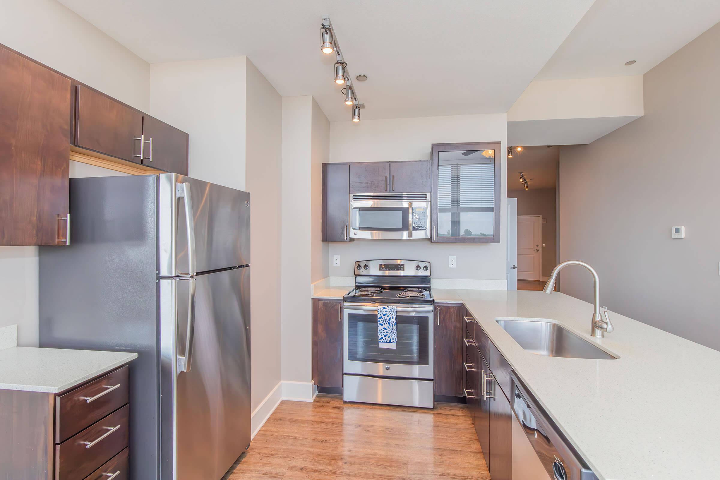 Modern kitchen featuring stainless steel appliances, including a refrigerator, oven, and microwave. The cabinetry is dark wood, contrasted with a light-colored countertop. Bright overhead lighting enhances the spacious feel, with an open layout leading to a well-lit area. Elegant design and functionality are key elements.