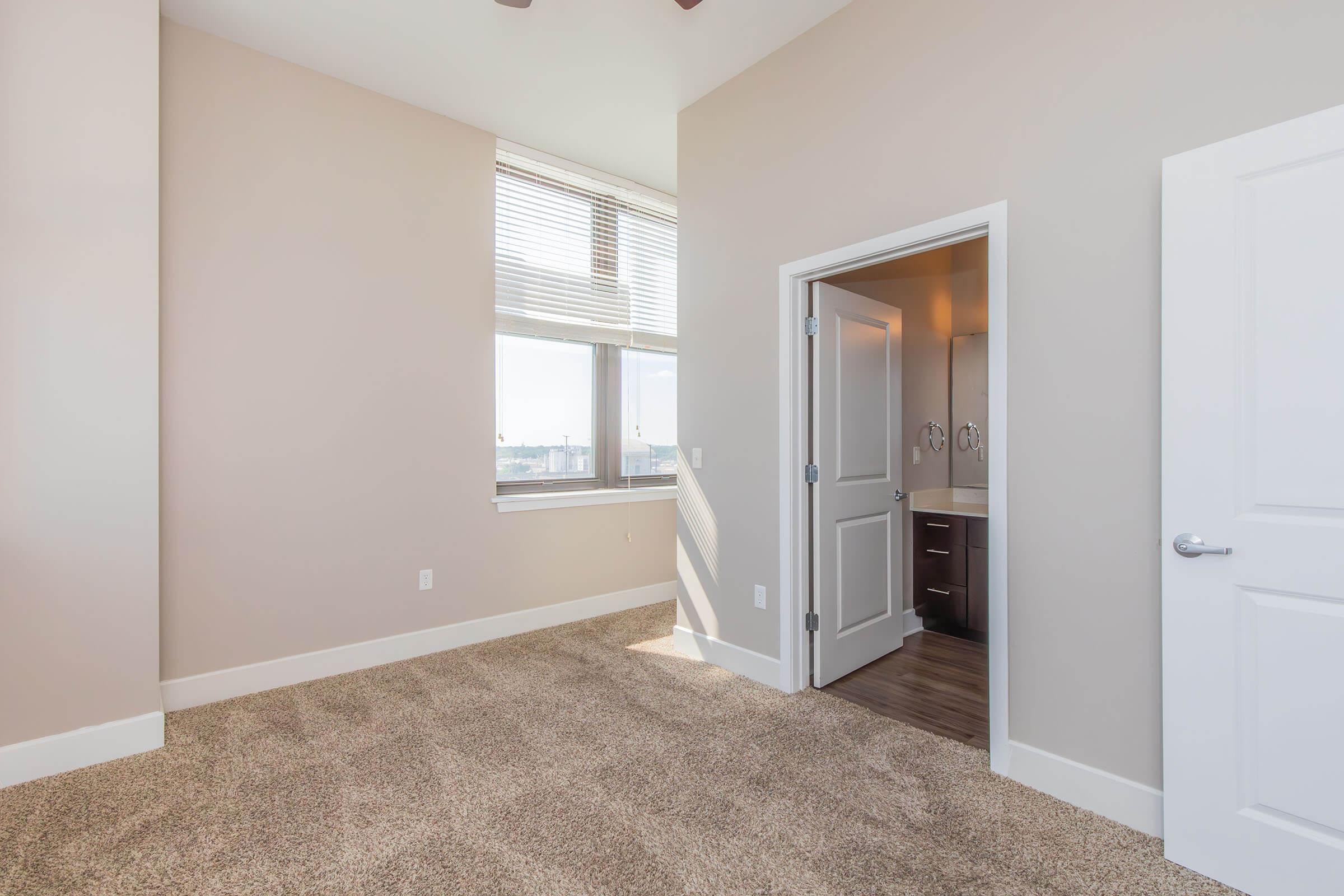 A well-lit, spacious room with neutral-colored walls and carpet. Large windows allow natural light to enter, and an open door leads to a modern bathroom. The space features a ceiling fan and a clean, minimalist design, creating a comfortable and inviting atmosphere.