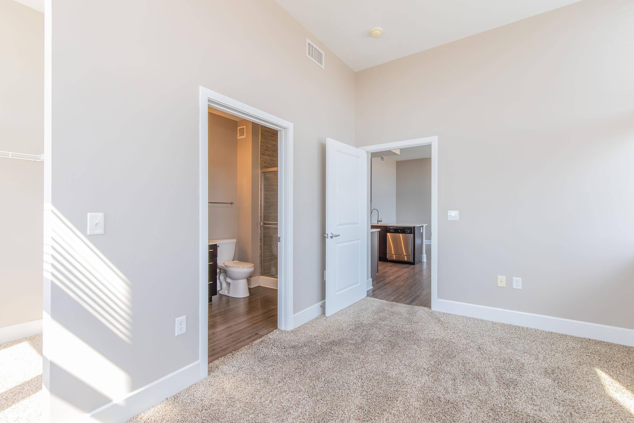 A spacious interior view of a room with light-colored walls and carpeted flooring. There are two white doors, one leading to a bathroom with a toilet and sink visible, and the other to a kitchen area with modern appliances. Bright natural light fills the space through a window.