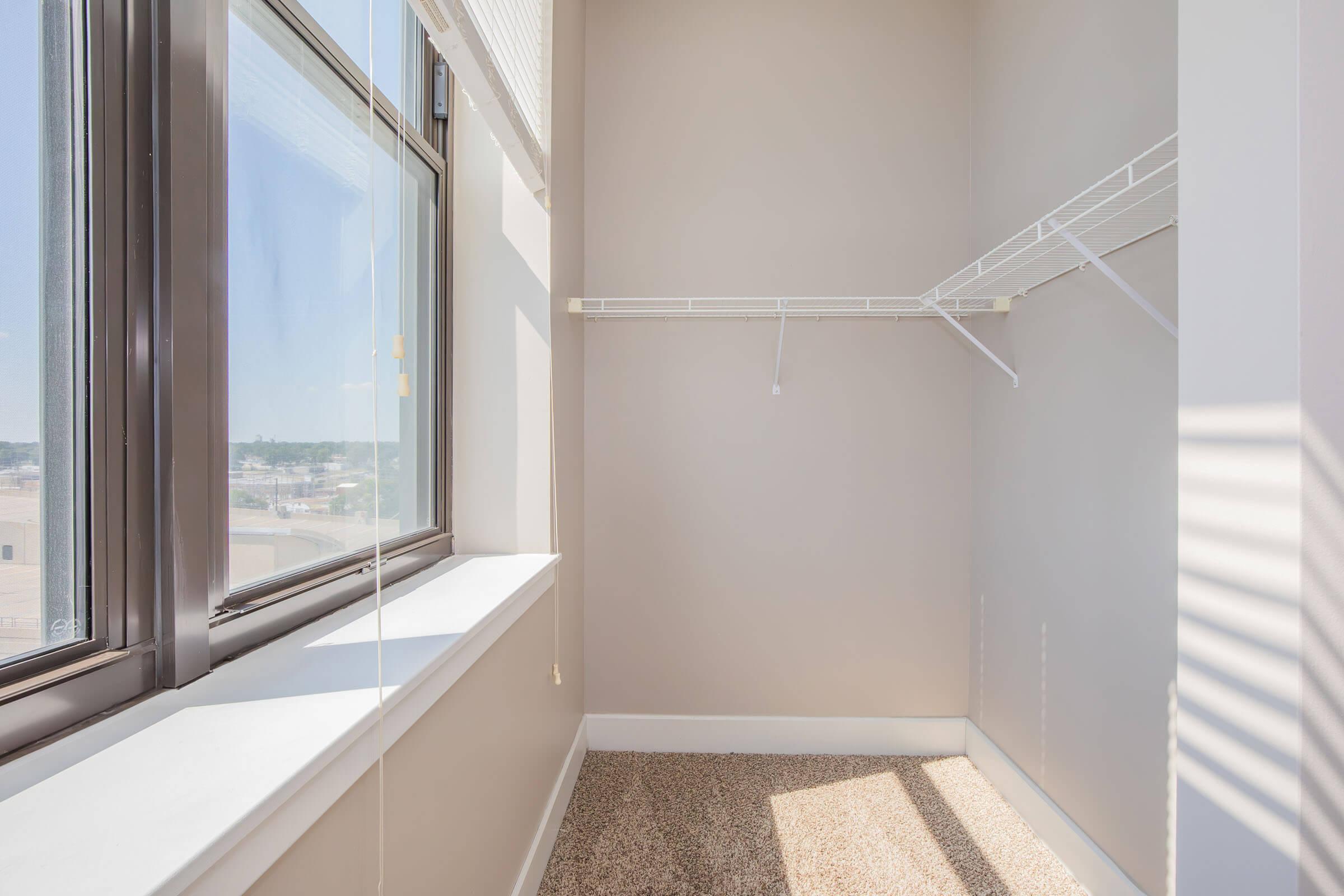 A light-filled corner of a room featuring a large window with blinds, showcasing a view of the outdoors. The interior has beige walls and carpeting, with a white wire shelving unit mounted on one wall. The space is empty, conveying a clean and modern feel.