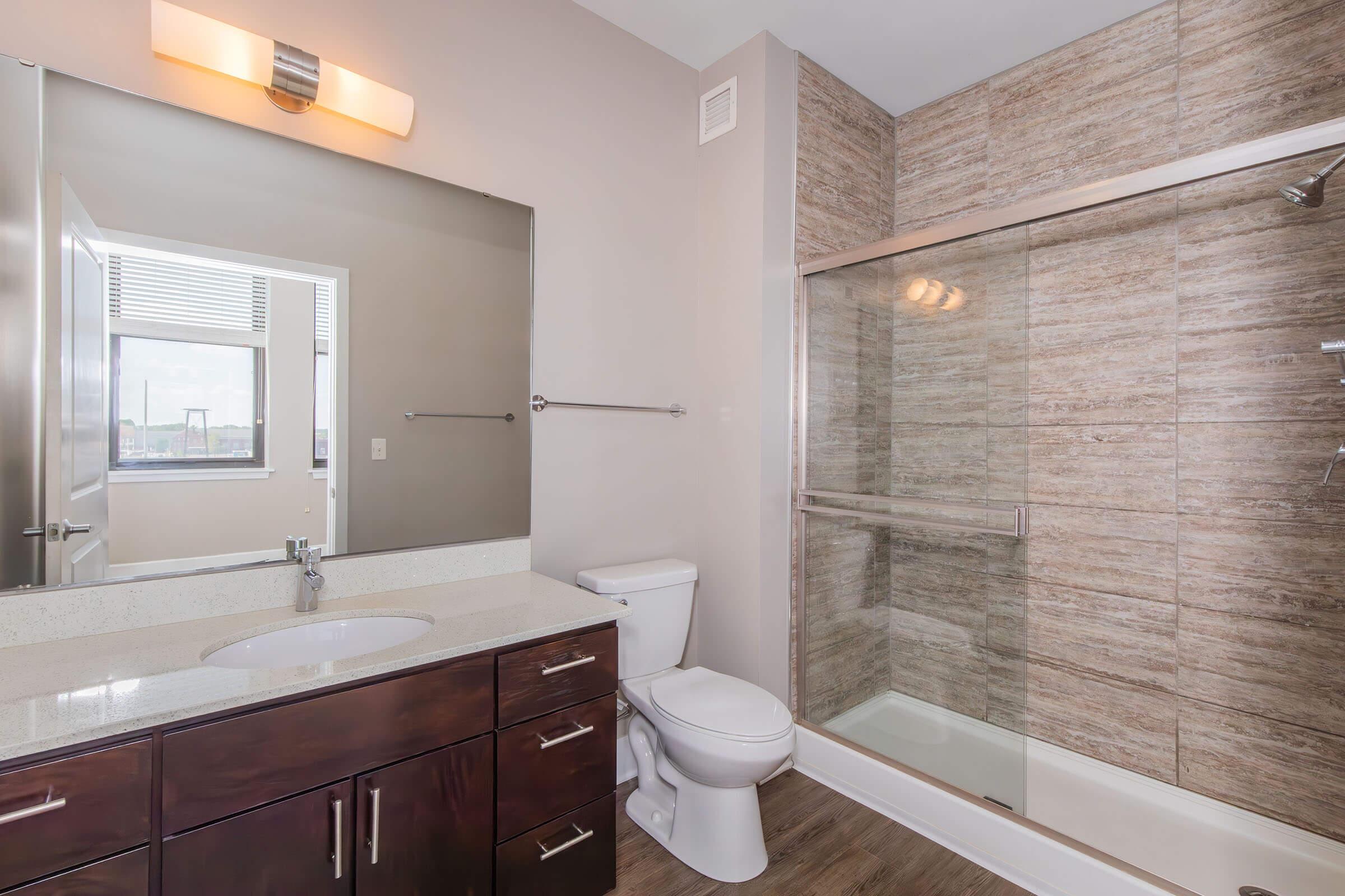 Modern bathroom featuring a glass shower enclosure, a white toilet, and a sleek vanity with a sink. Neutral-toned walls and a spacious mirror enhance the bright atmosphere, while wood-like flooring adds warmth to the design.