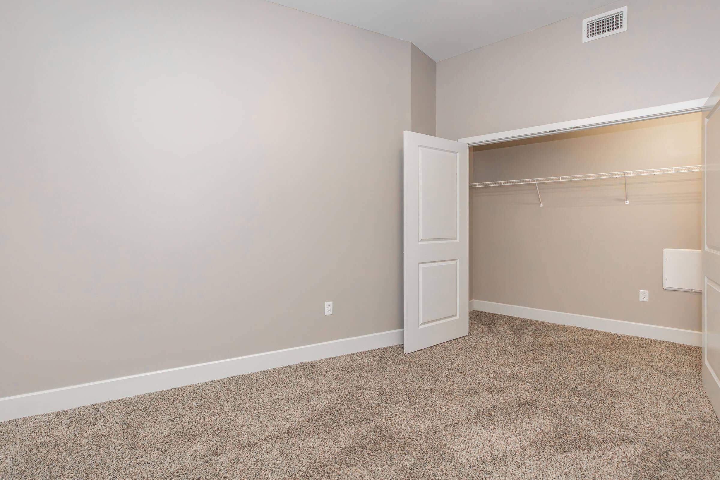 A spacious, empty closet with light-colored walls and a light carpet. The closet has a white door slightly ajar, revealing a hanging rod and shelves inside. The room appears well-lit and modern, with minimal decor.