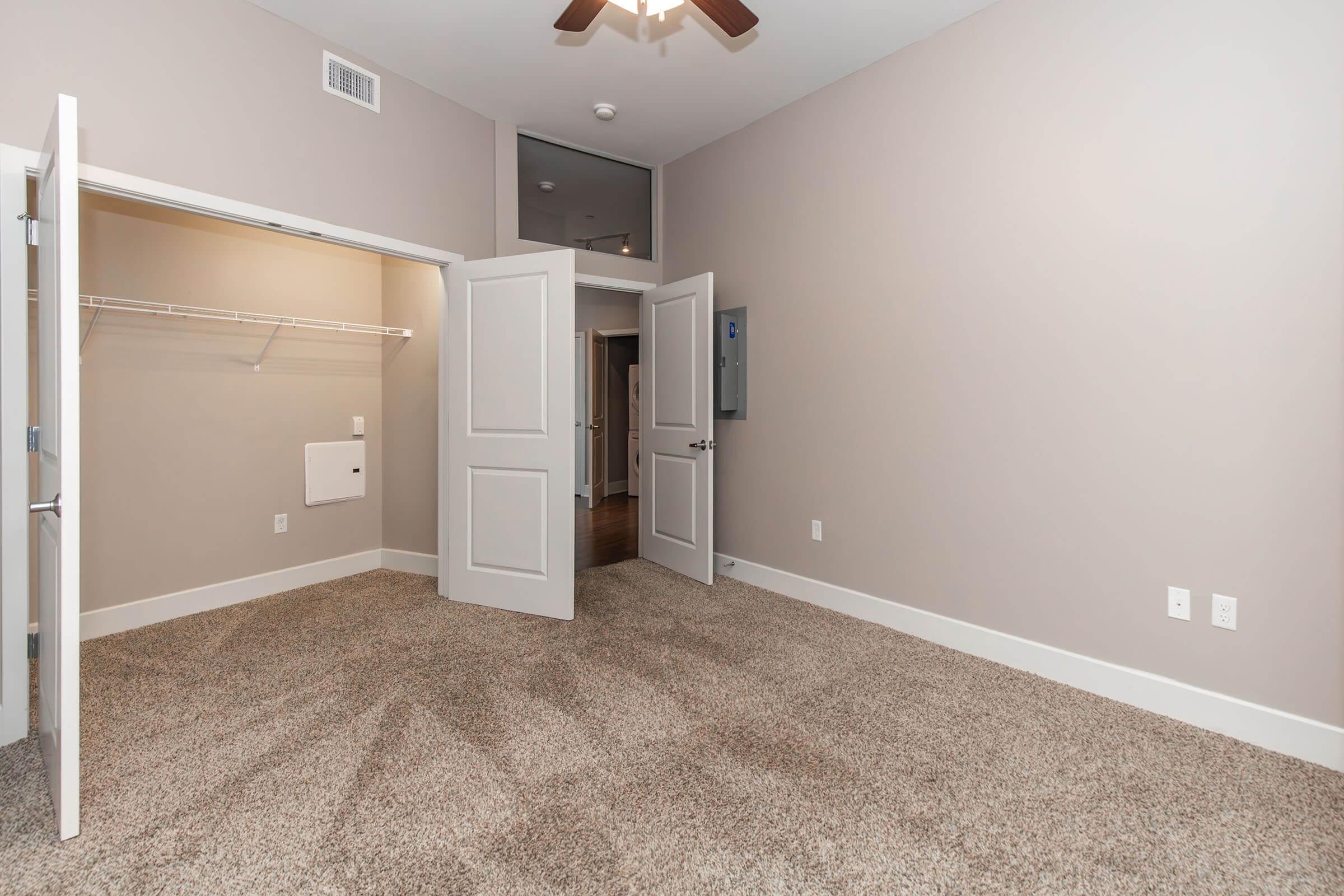 A spacious room with light-colored walls and carpeted flooring. Two open double doors lead to a closet on the left. A ceiling fan is visible, and a wall-mounted electrical panel is on the right. The room has a clean and modern look, with natural light illuminating the space.