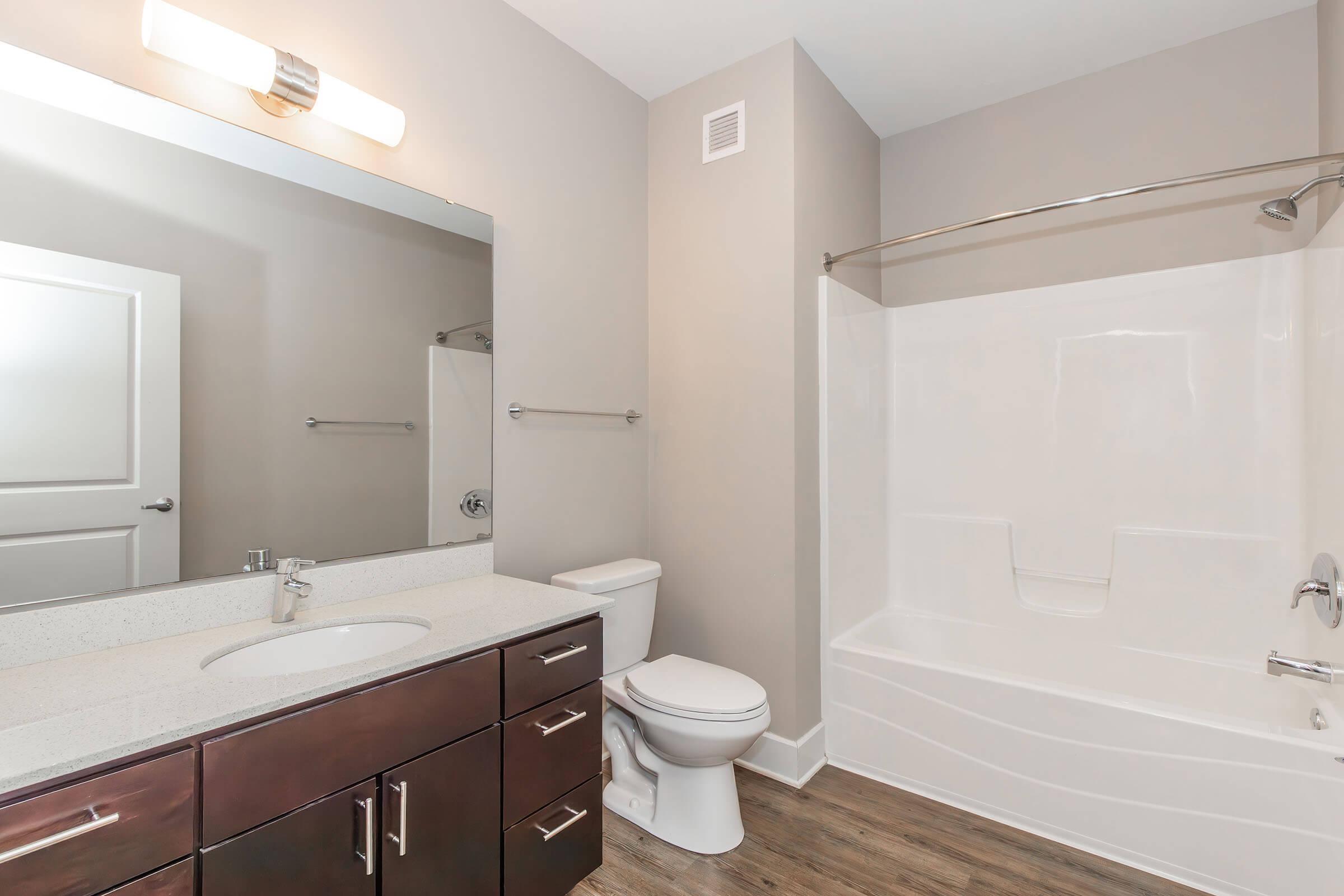 A modern bathroom featuring a large mirror above a white sink with a granite countertop, dark wood cabinetry, a white toilet, and a bathtub with a shower. The walls are painted in a light gray color, and the flooring has a wood-like finish. A soft overhead light illuminates the space.