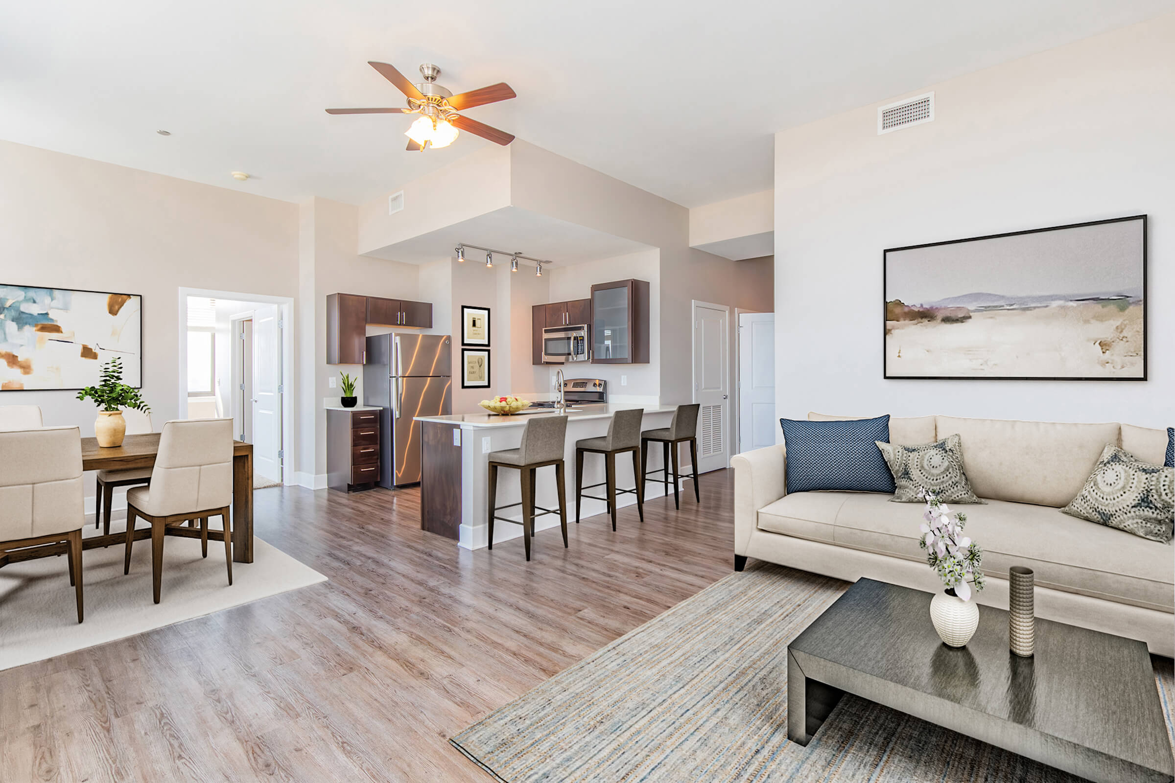 Modern living room and kitchen area featuring light-colored walls, hardwood flooring, and contemporary furniture. The living room has a beige couch with cushions, a coffee table, and a decorative flower vase. The kitchen showcases stainless steel appliances and bar stools at a counter. Natural light flows in through large windows.