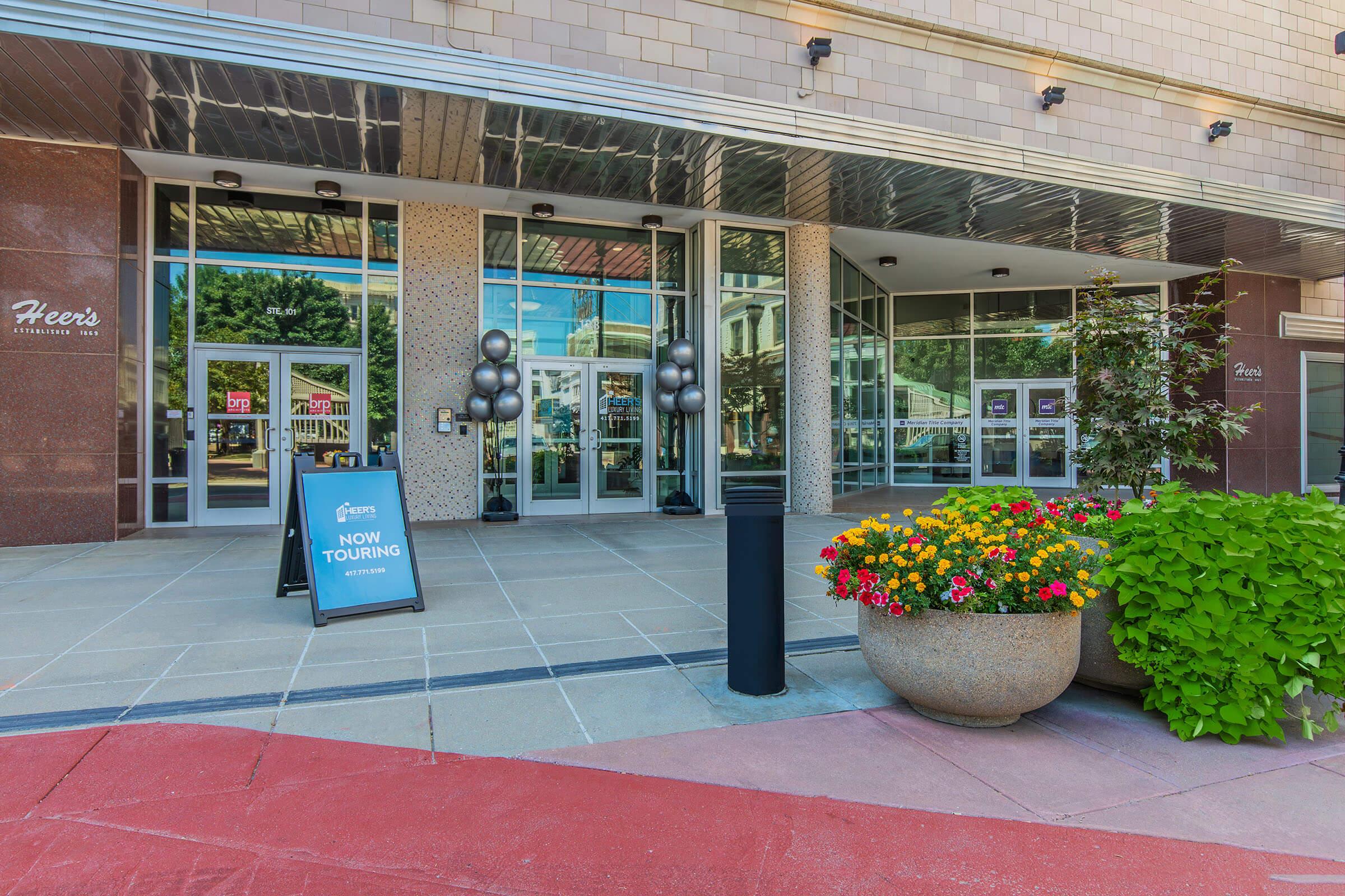 Entrance to a modern building featuring large glass doors, a sign that reads "NOW TOURING," and colorful flower planters. The exterior has a polished stone finish and decorative elements, with a bright walkway leading up to the entrance. The surrounding area includes trees and a clear blue sky.