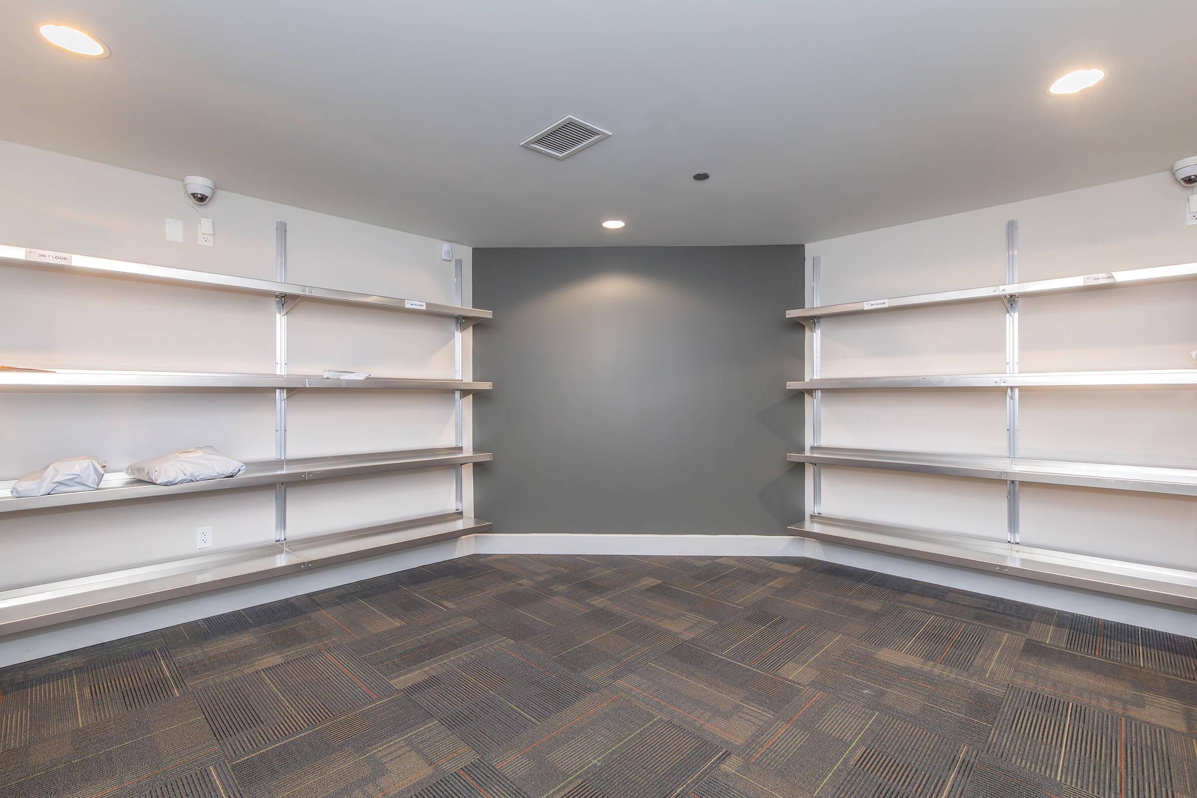 A small, empty room featuring two walls with silver shelving units mounted. The shelves are sparsely populated with a couple of pillows, while the floor is covered with gray carpeting. Ceiling lights illuminate the space, and security cameras are visible in the corners. The walls are painted a neutral color.