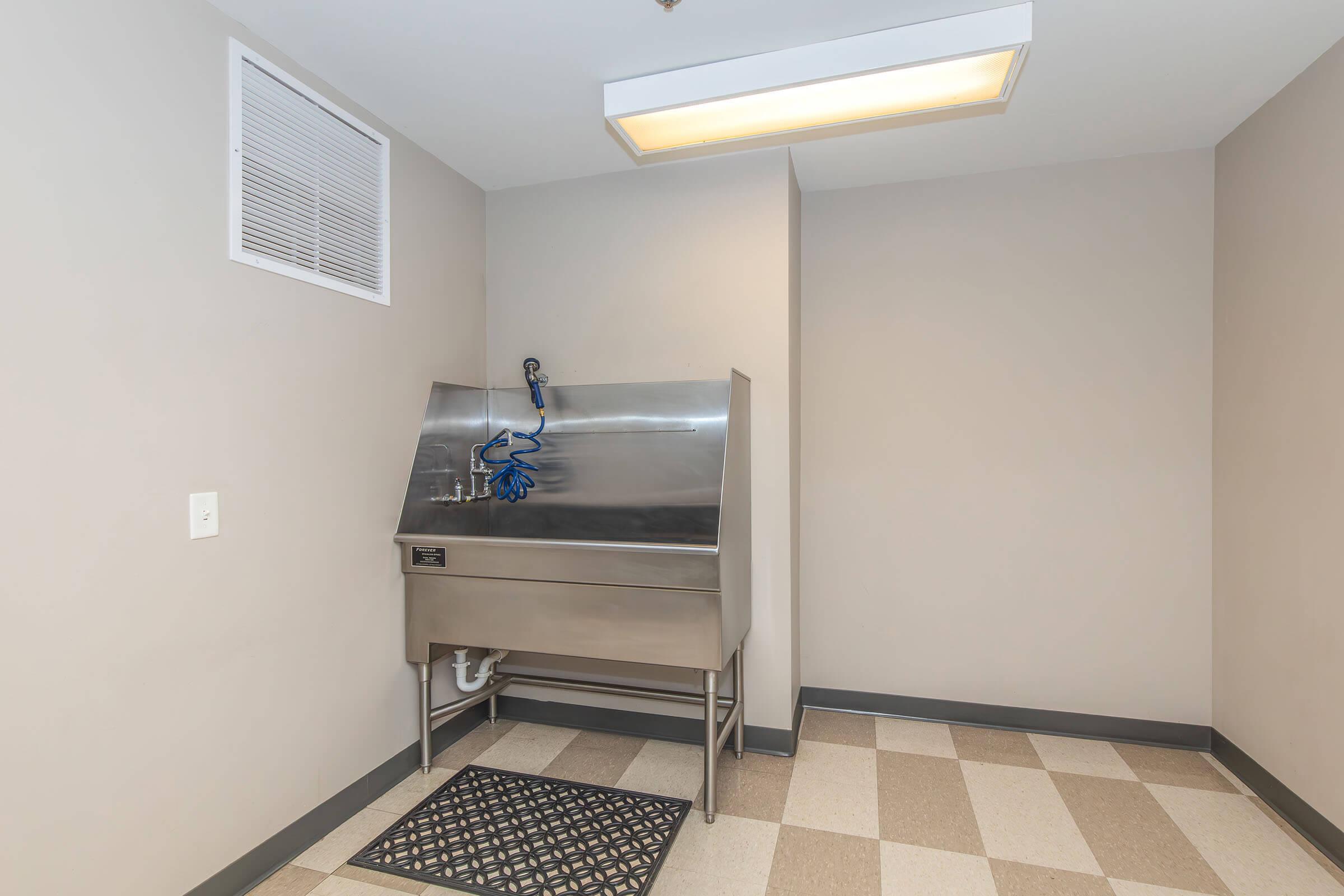 Stainless steel utility sink located in a room with light gray walls and checkered floor tiles. The sink features a blue hose attachment and is positioned against a wall with a ventilation vent above it. A black rubber mat is placed in front of the sink.