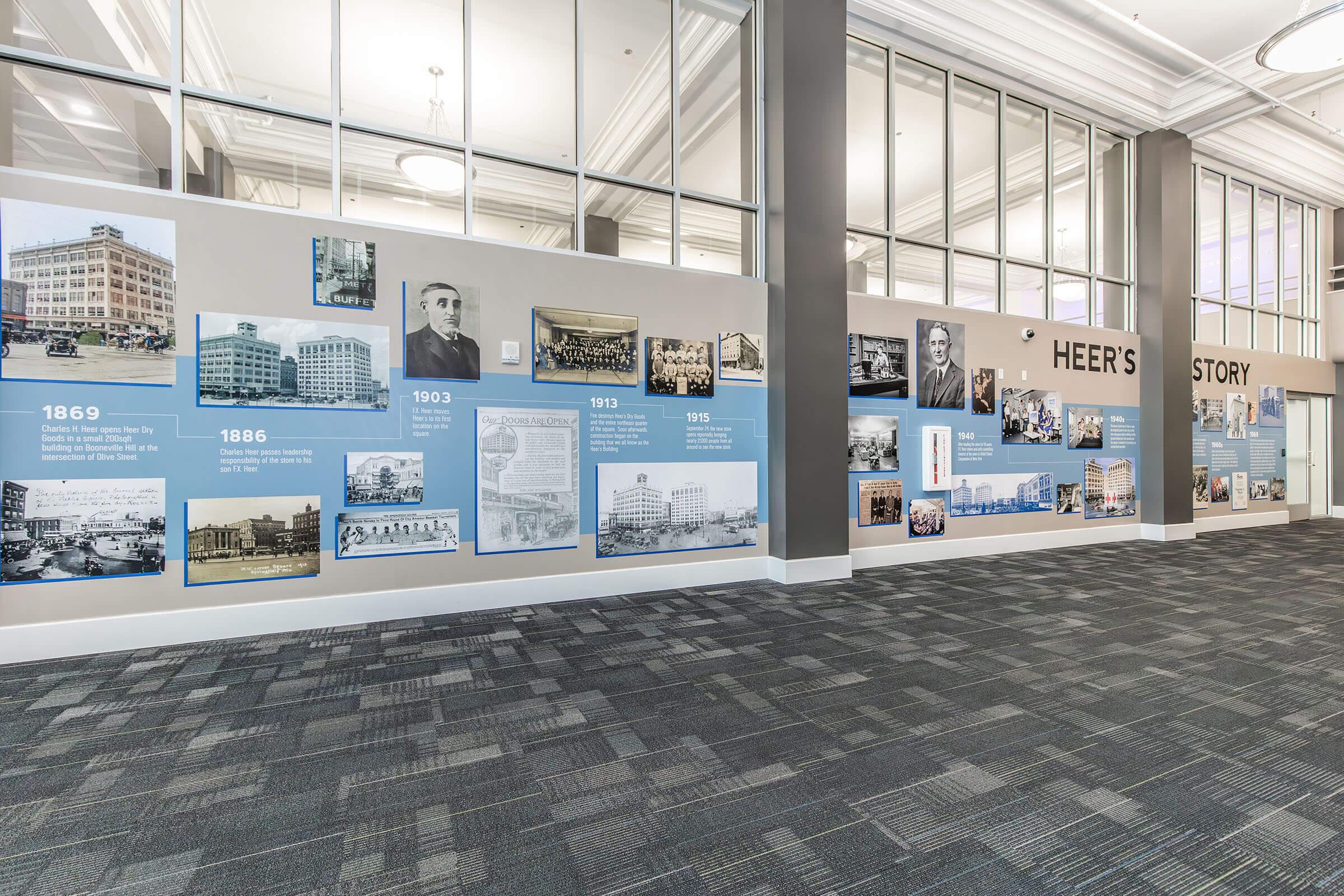 A modern exhibition space featuring a wall displaying historical photographs and text panels that narrate the timeline and story of Heer’s, including significant years like 1869, 1886, and 1903, in a well-lit area with a carpeted floor.